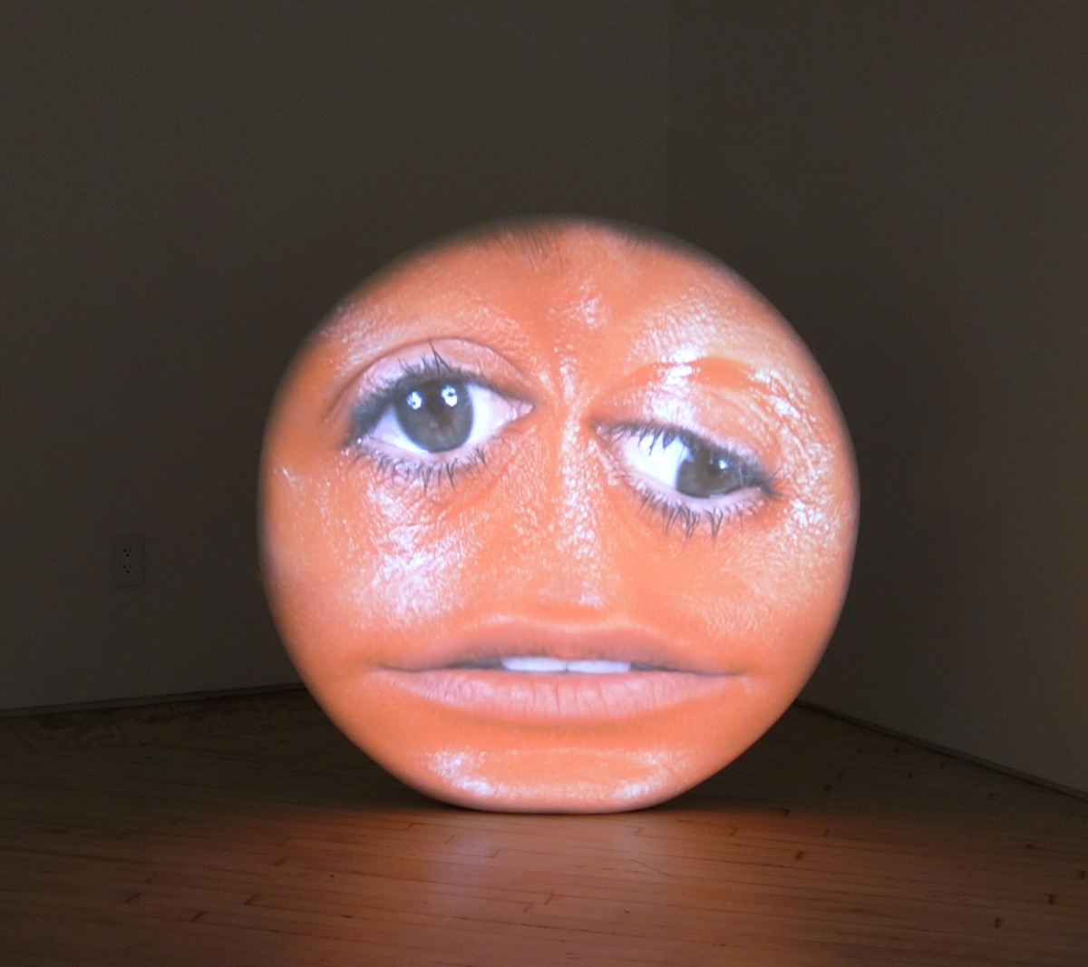 Tony Oursler, pEachhhh?, 2004, Projection on foam and resin sclupture, audio - round object in a darkened room, a round, close up head painted red is projected on the surface. The face has two eyes and a mouth, no nose.
