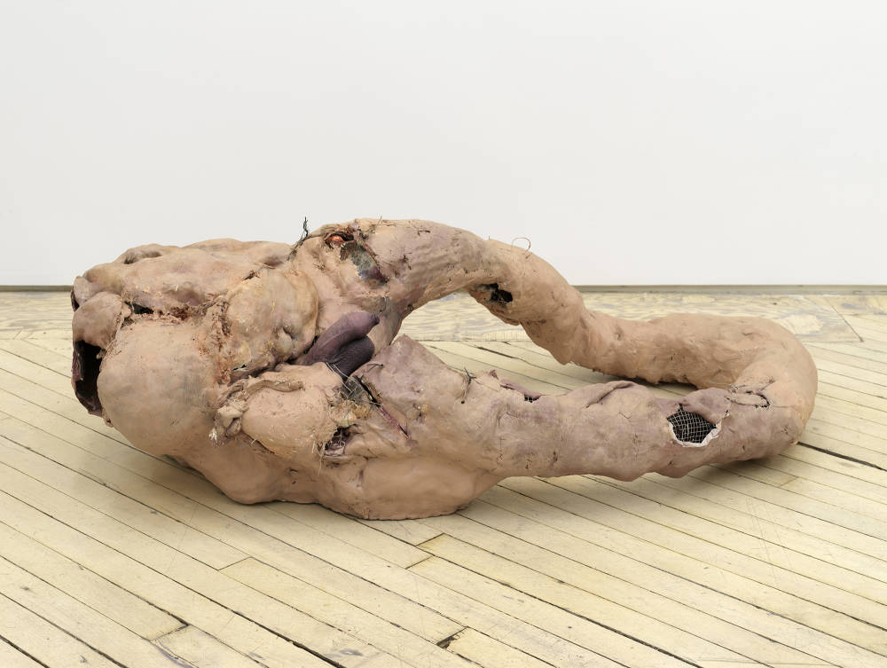 In a gallery space, an abstract sculpture rests on the floor resembling human flesh.