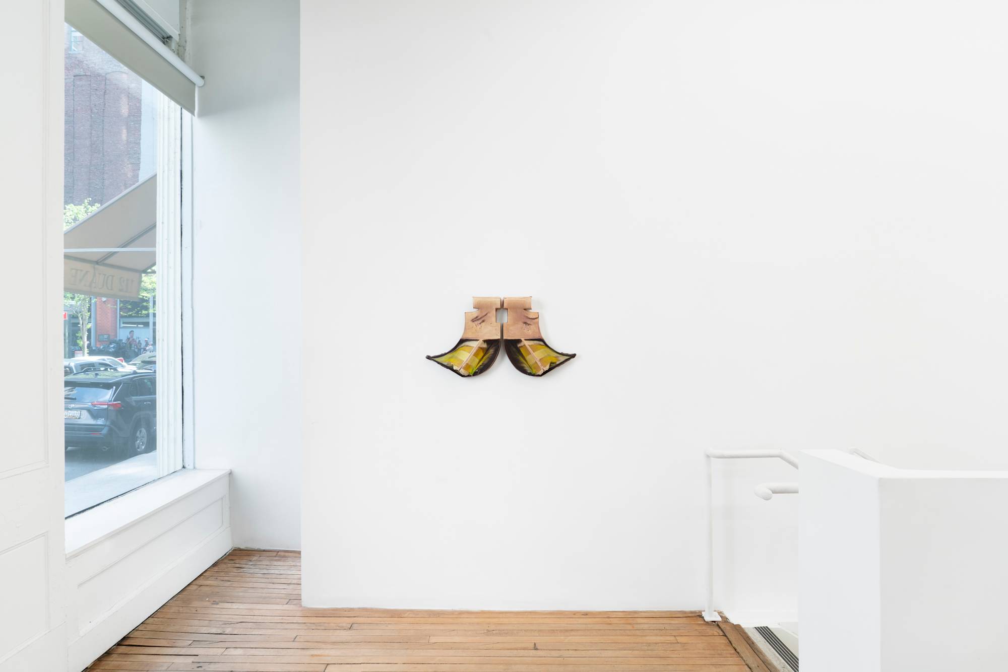 Two ceramic sculptures hanging on a wall of a gallery with wood floors. The two pieces resemble the letter "J", one forwards, one backwards, mirroring each other. A spine runs diagonally down each bottom swoop, striped with yellow and green glaze, against a background of dark brown glaze. A large window to the street is visible on the wall to the left and a staircase descends to the right out of frame.