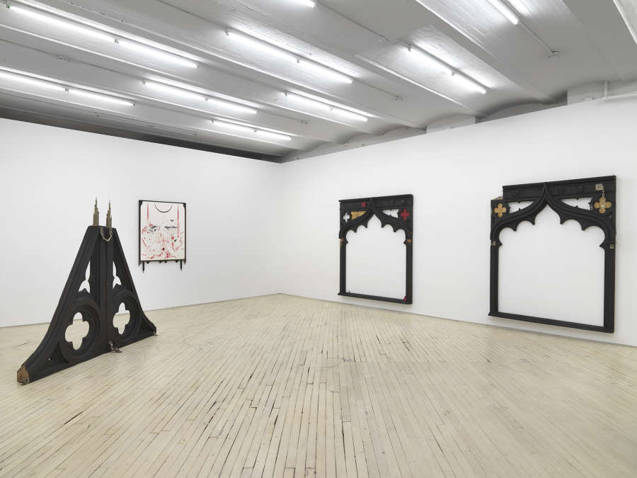 Installation view of main gallery: at left, a pyramid shaped gothic style wooden floor sculpture behind which is a black and red drawing hanging on the center wall, framed in black wood, at right two large wooden archway forms hangs on the wall.