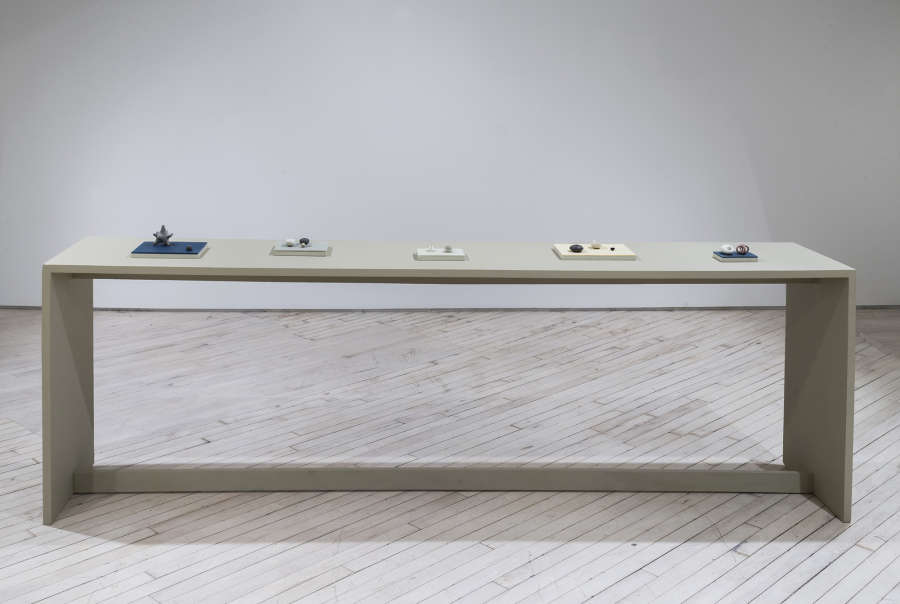 In a gallery space, four miniature arrangements of abstract sculptures are placed on small colorful plinths spaced evenly apart on a green-tinted table. 