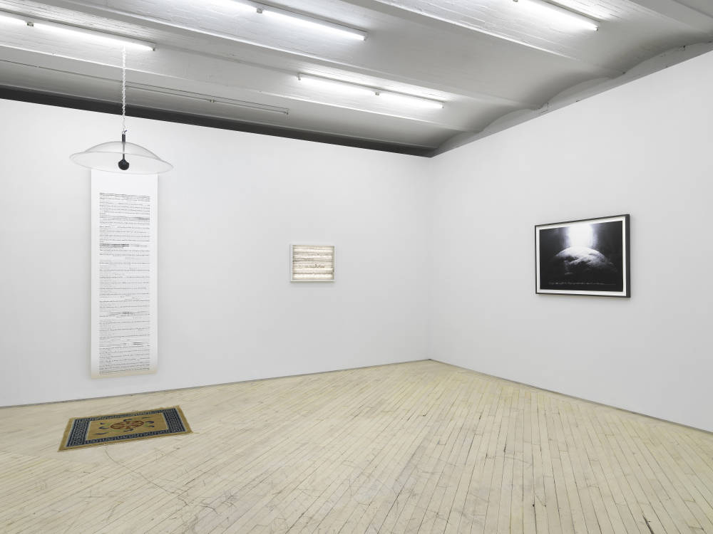 In a gallery space, three wall mounted artworks hung evenly spaced apart. To the left a long scroll with a parabolic speaker hanging in front of it above a rug. 