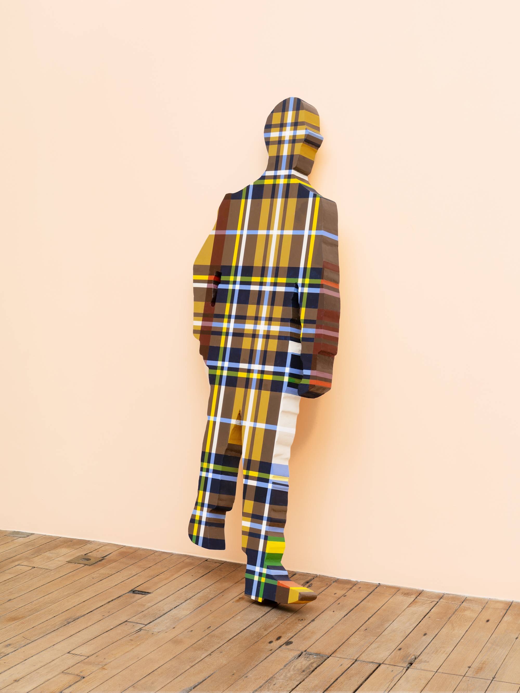 A life-sized human figure painted in a brown, yellow and blue plaid pattern leans against a peach wall in an art gallery.