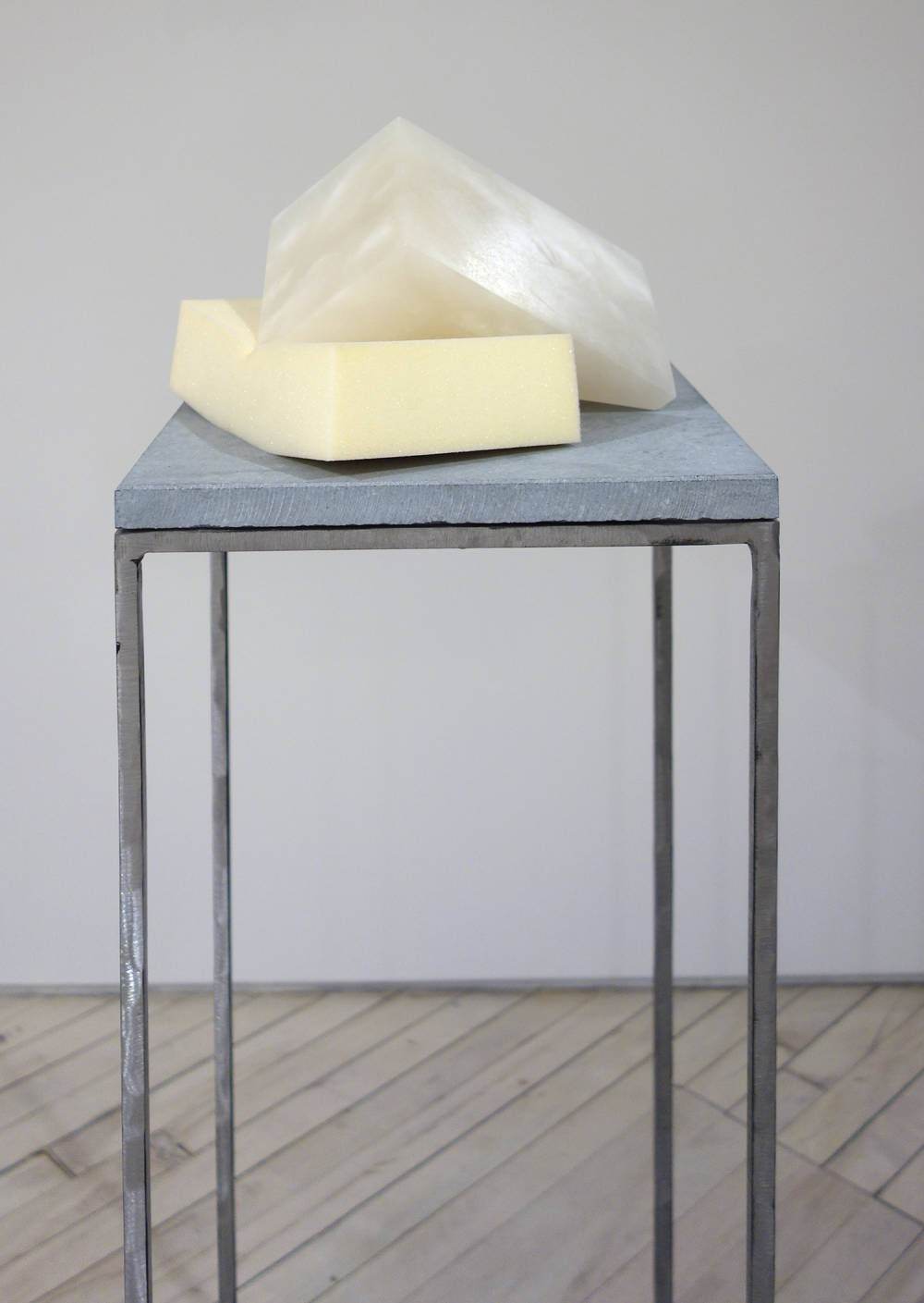 Atop a blue stone and steel plinth sits a rectangular block of white alabaster. There is a triangular piece of the alabaster cut from the block, and it is resting on its side atop block of memory foam pressing into the foam.