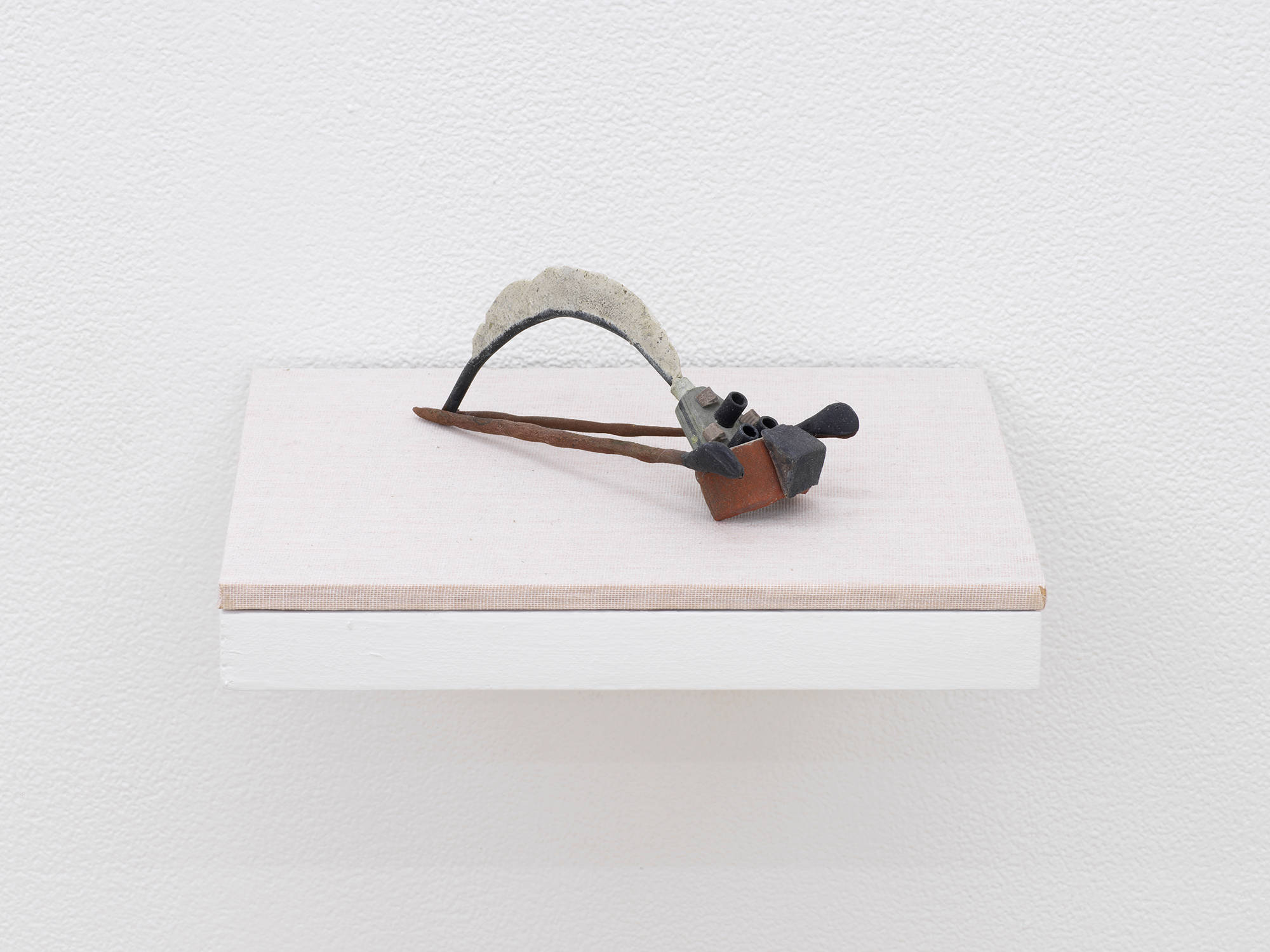 A small abstract sculpture resembling a kind of prehistoric tool or instrument. The object is primarily brown, black and gray and resting on a shelf with a tan base attached to a white wall.