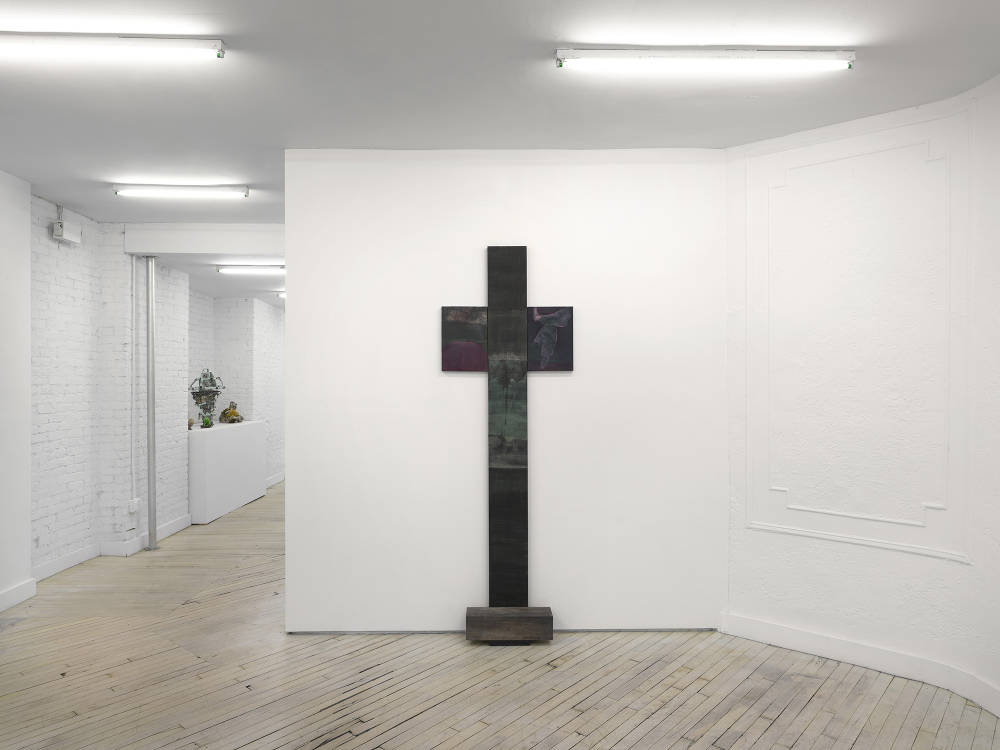 A vertical sculpture leaning on a wall in the shape of cross leaning against a gallery wall. 