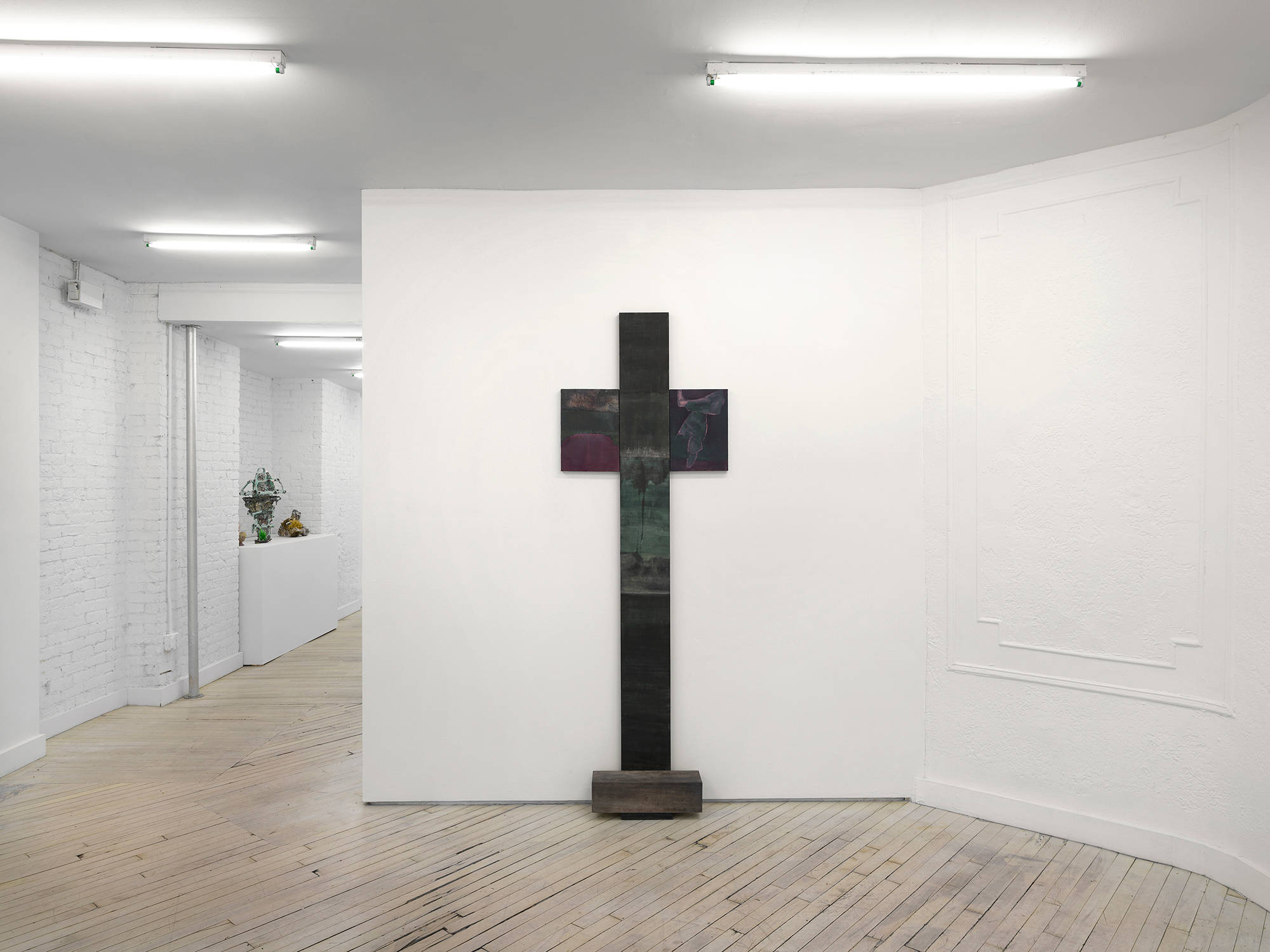 A vertical sculpture leaning on a wall in the shape of cross leaning against a gallery wall.