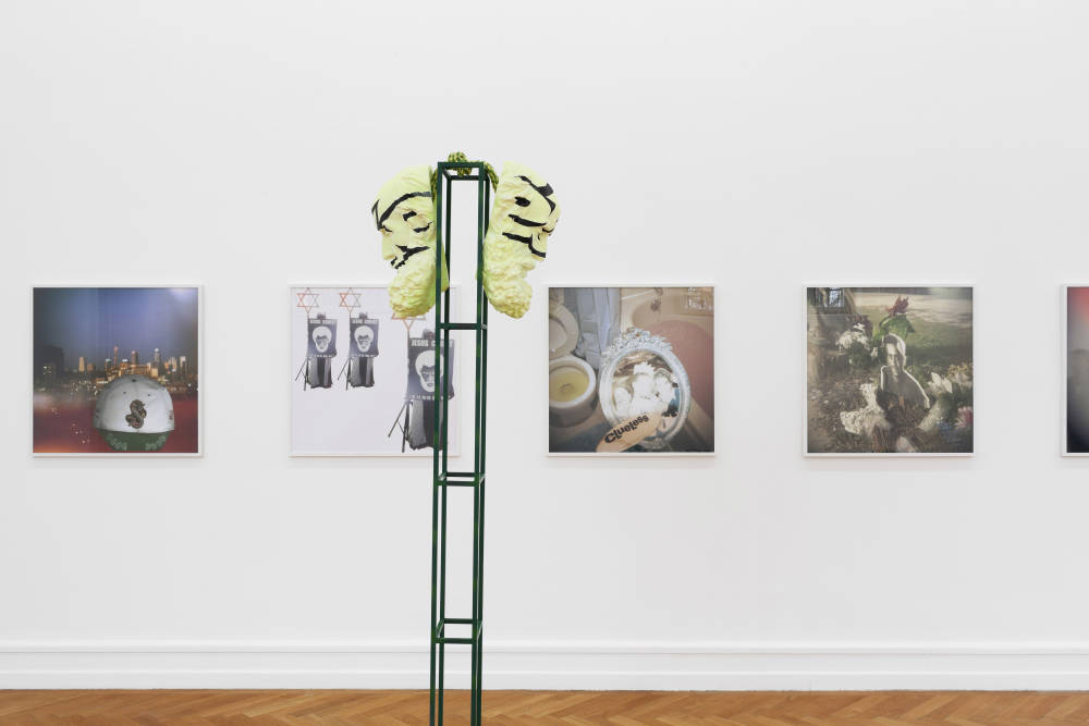 In a gallery space five framed photographed in white frames capturing vague interior and exterior 
spaces. In the foreground a tall steel structure with two abstract forms hanging from either side of the top of the structure. 