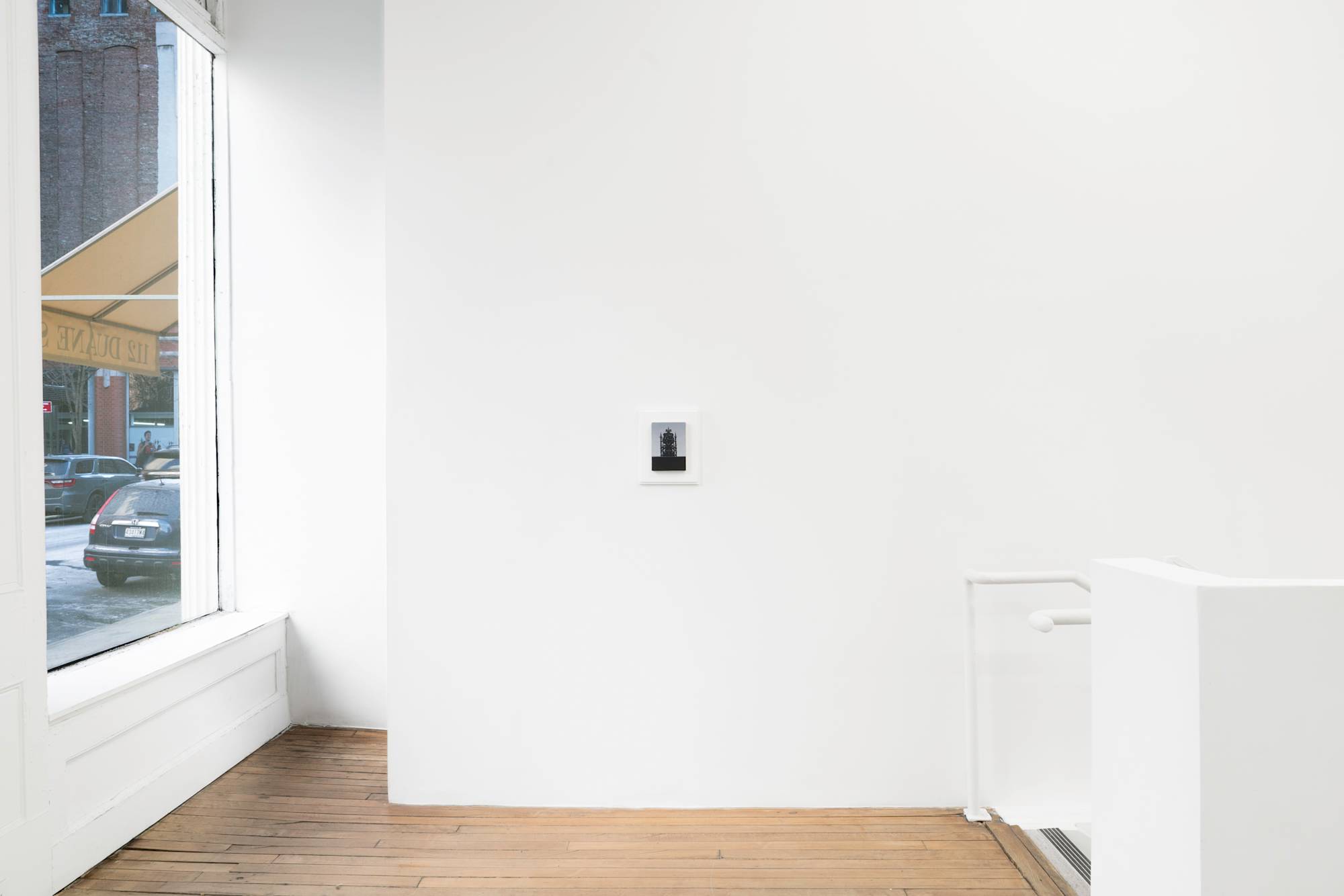 Ian Miyamura painting depicting a throne of skulls against a light grey background hangs on the wall of a gallery. The painting is mounted to a white frame made by the artist. A staircase leads downstairs to the right of frame.