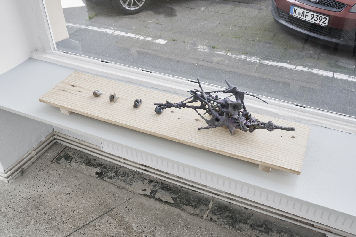 Four small hand-sized sculptures and one medium scale sculpture are presented on a rectangular piece of wood in front of a large window that opens up to a street view. The sculptures are amorphous, skeletal, and gray. They are resting on a bench inserted between two walls. 