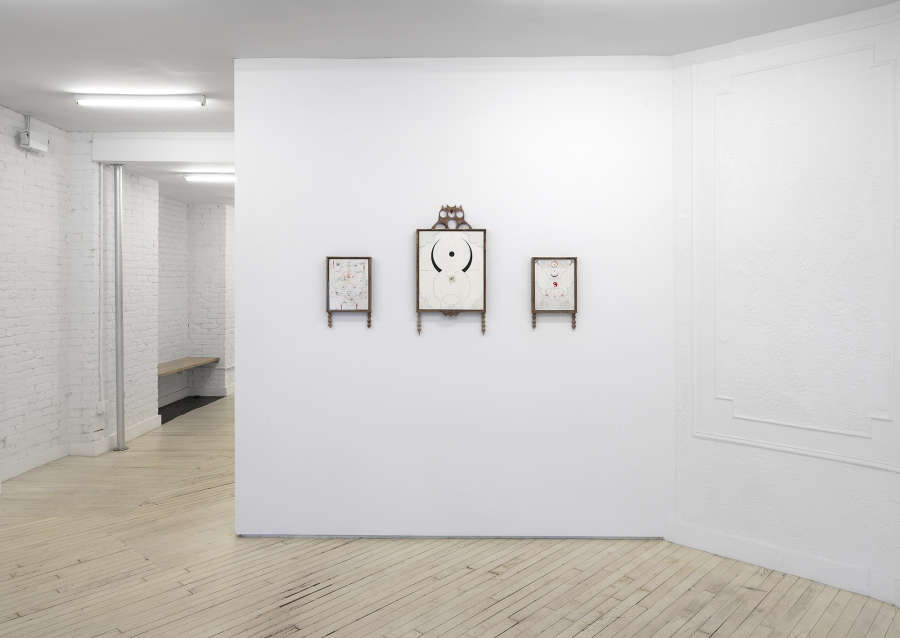 Image of three framed drawings, two small drawings flanking one larger drawing, all framed in wood and hanging on a white wall with a hallway on the left side of the image. 