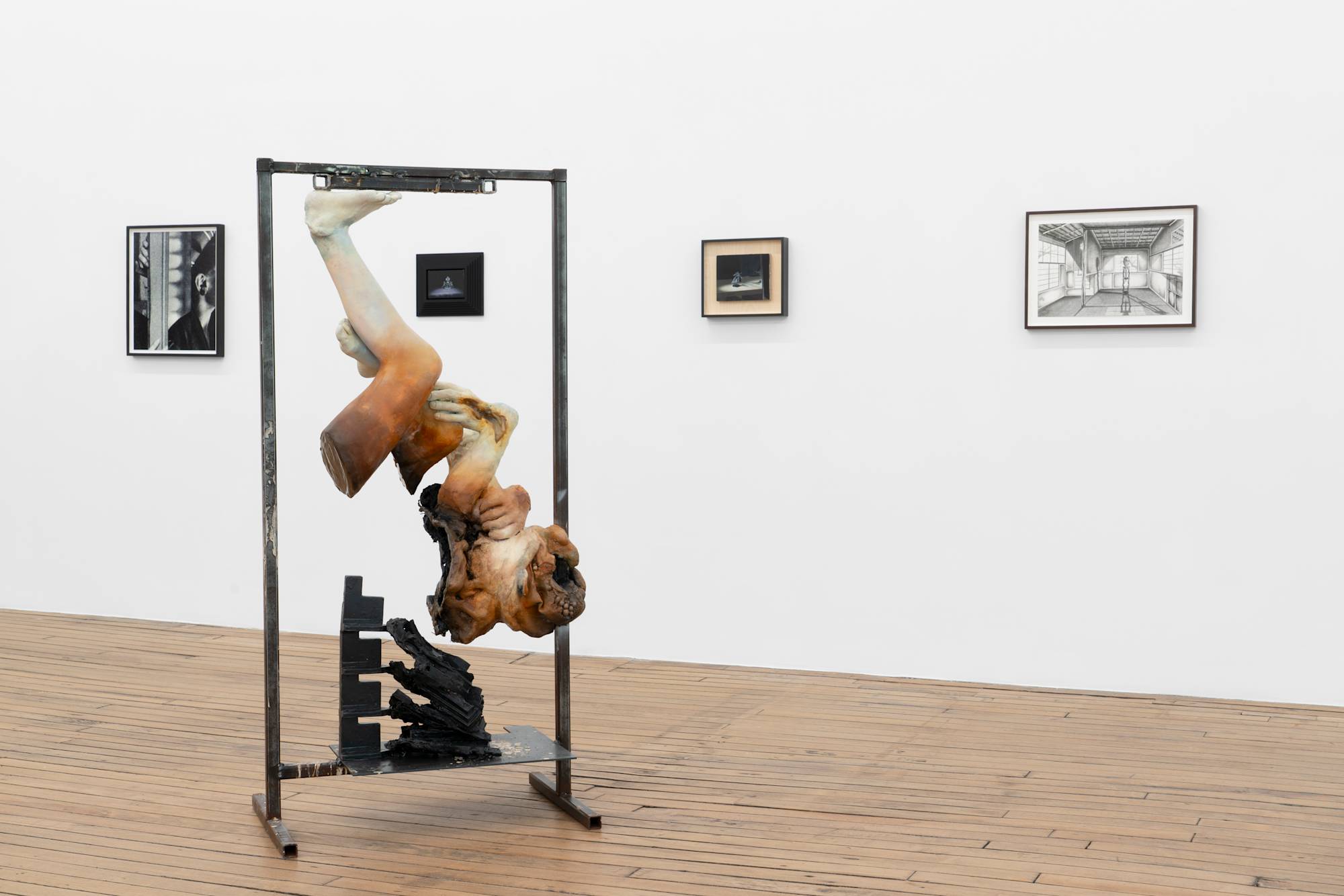 A freestanding sculpture featuring a fragmented figure, inverted and suspended from a metal frame, stands on the wooden floor of an art gallery. Four smaller framed works of art are displayed on the walls surrounding the sculpture.