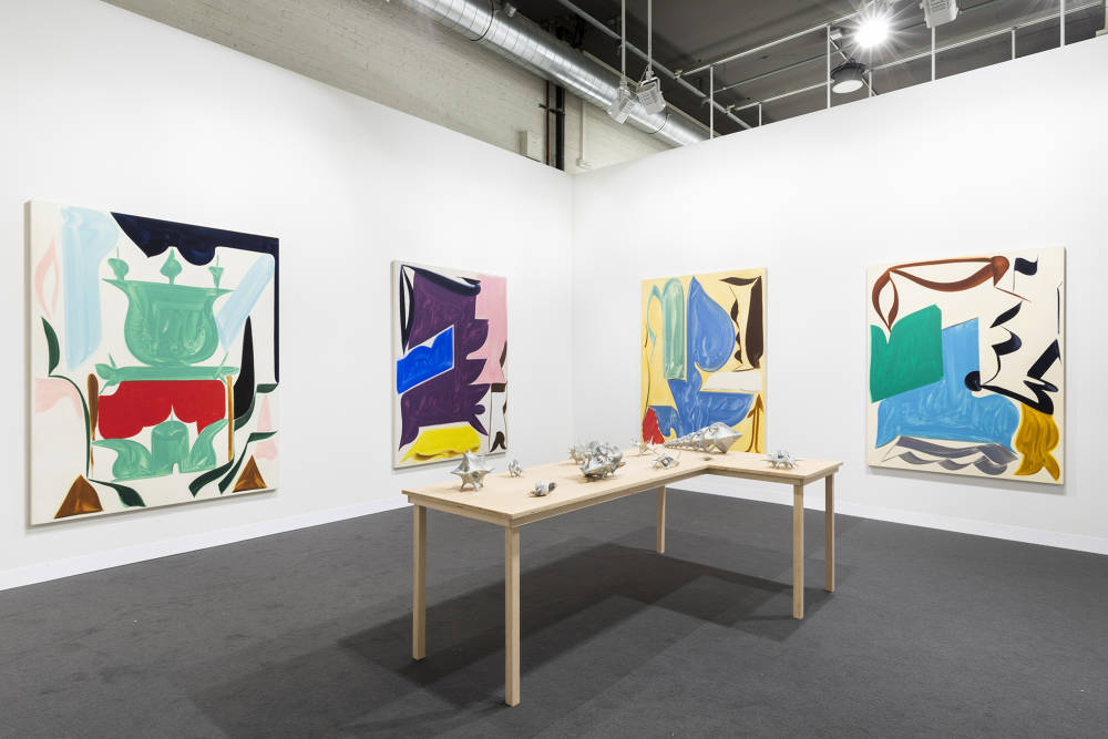 In a gallery space, four large abstract paintings are spaced apart. The painting depict bold abstract geometric shapes in various hues of color. The dominant colors are hues of yellow, purple, pink, blue, green, brown, and black. The paint is thin and gestural. In the center of the gallery space is a freestanding brownish table with various miniature gray sculptures placed evenly apart. The sculptures are abstract and resemble organic material or bone formations. 