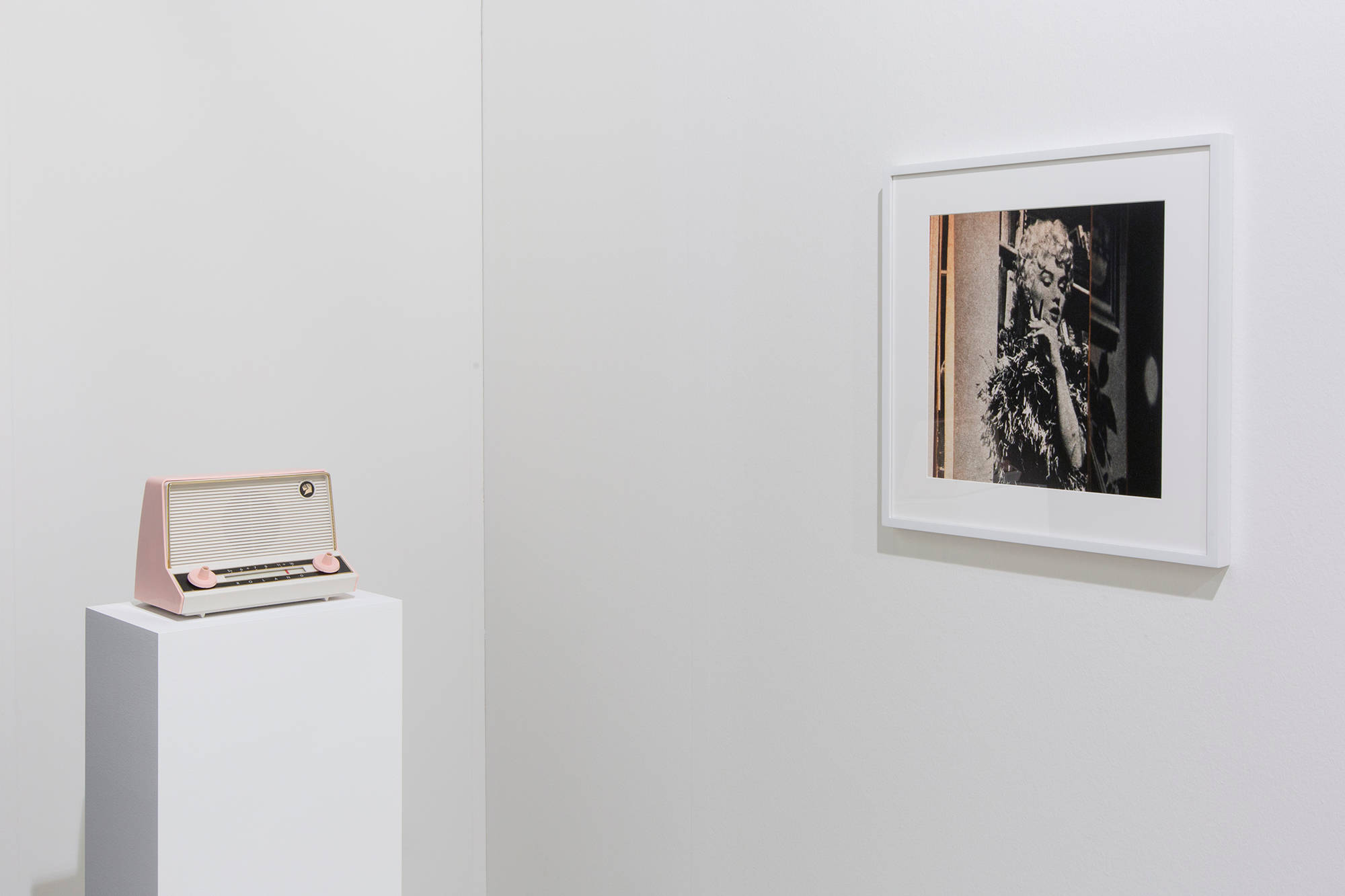Installation view; on the left is a white plinth with a vintage plastic radio which is pink and white. On the right is a color photograph of a book which is open to a page showing a black and white photo of Marilyn Monroe in a boa and smoking a cigarette.