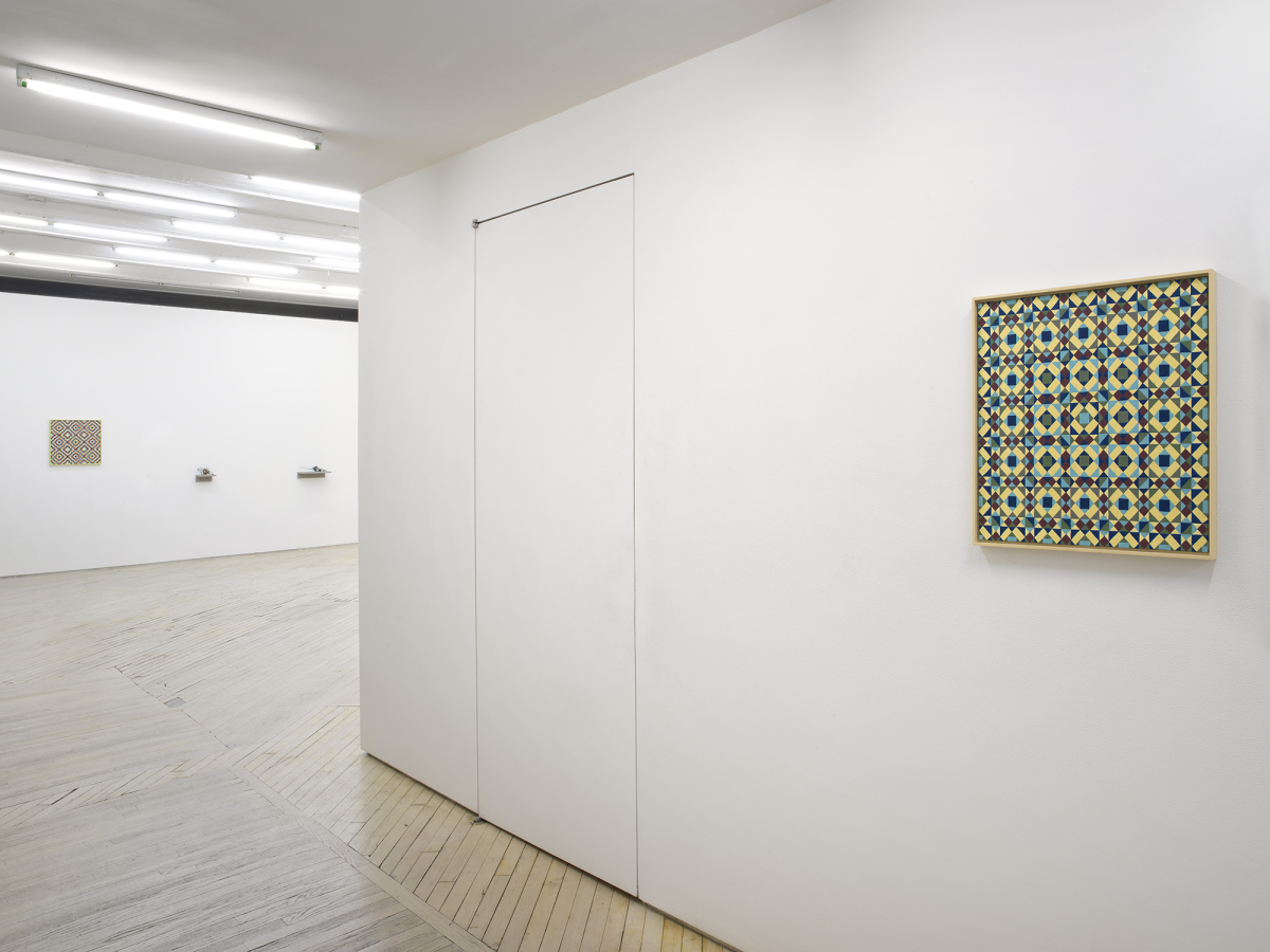 In the hallway of a gallery space, a painting depicting a repeating geometric pattern of criss-crossing lines. The dominant colors are hues of greens and blues. The frame is painted yellow. In the background is a larger gallery space with a similar painting hung to the left on the back wall. To the right of the paintings are two small sculptures installed on shelves lower on the wall. 