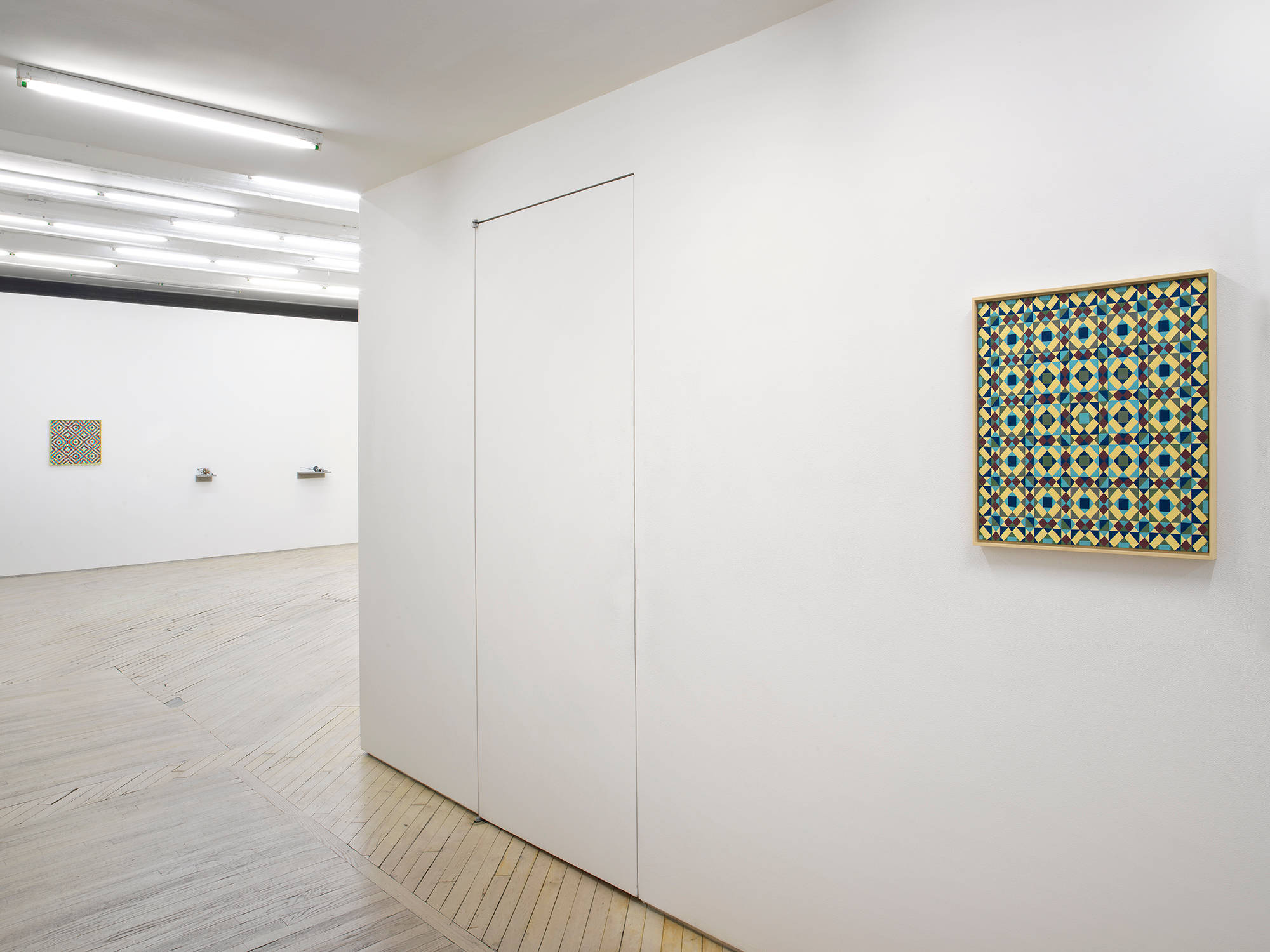 In the hallway of a gallery space, a painting depicting a repeating geometric pattern of criss-crossing lines. The dominant colors are hues of greens and blues. The frame is painted yellow. In the background is a larger gallery space with a similar painting hung to the left on the back wall. To the right of the paintings are two small sculptures installed on shelves lower on the wall.