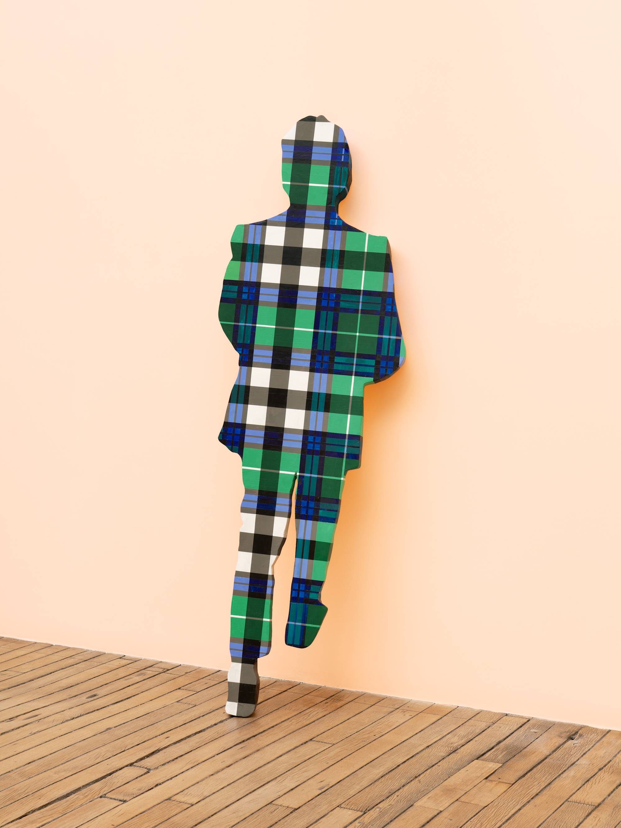 A life-sized human figure painted in a blue and green plaid pattern leans against a peach wall in an art gallery.