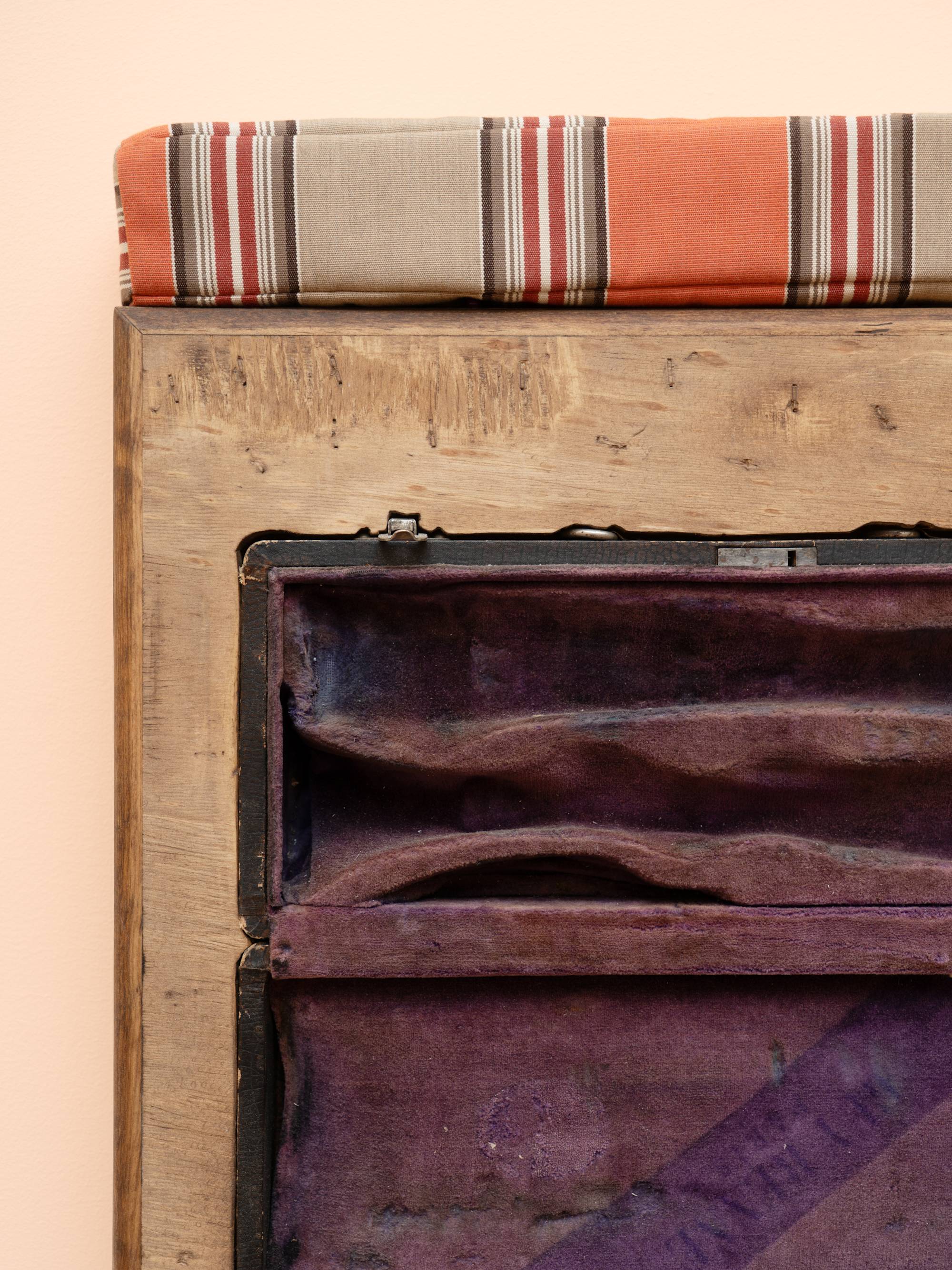 Detail view of a wooden box sculpture with inset clarinet case with purple velvet lining, topped with a orange and beige striped cushion, hanging on a peach colored wall.