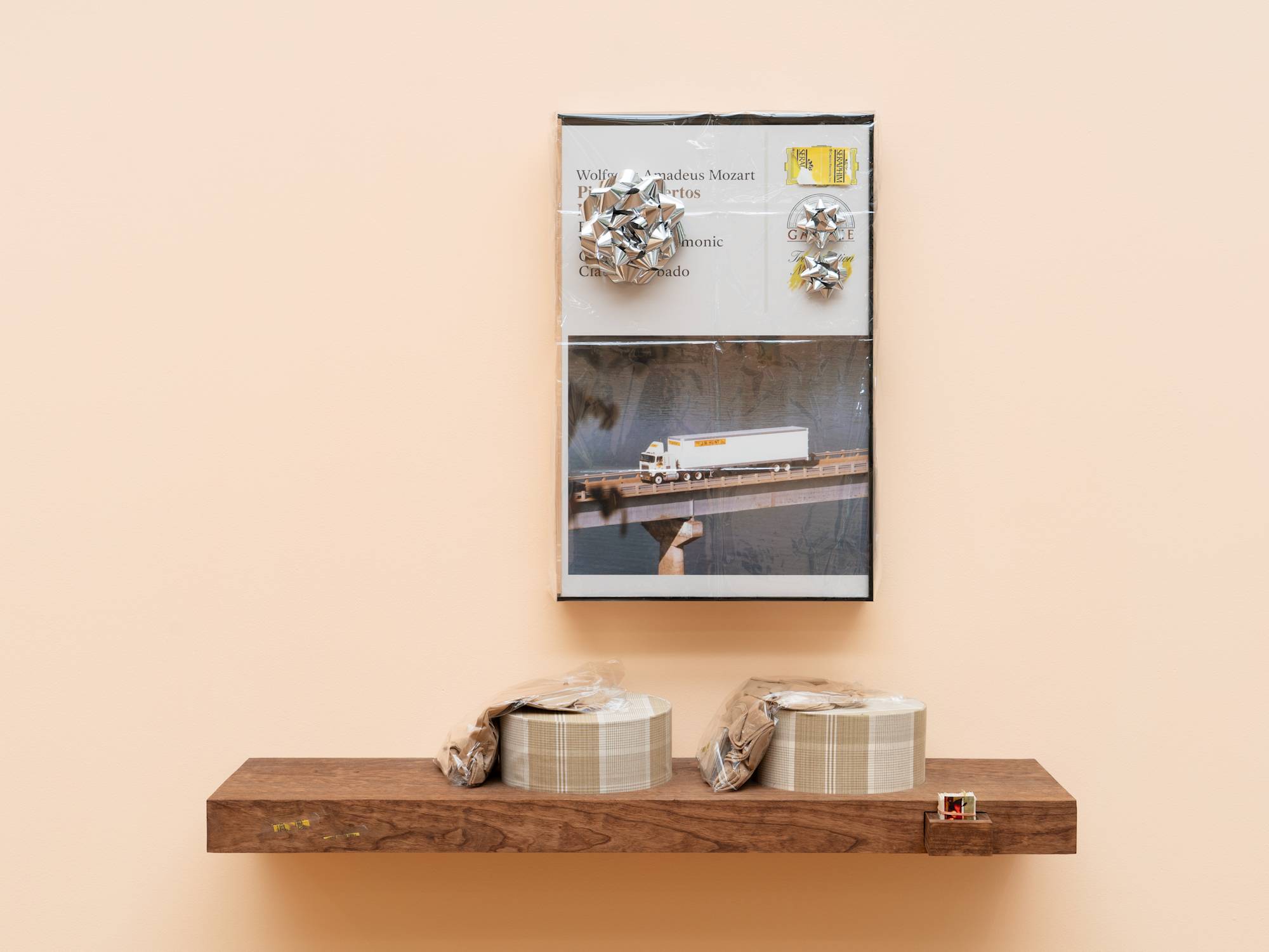 A wood veneered shelf hangs on a peach colored wall. 2 tan, plaid, circular gift boxes sit on the shelf with tan socks in plastic bags draped over them. A small drawer juts out of the front of the shelf and is neatly filled with trimmed business cards. A large, fake audio cassette case featuring a truck driving over a bridge hangs above the shelf, wrapped in cellophane and decorated with three silver gift bows.