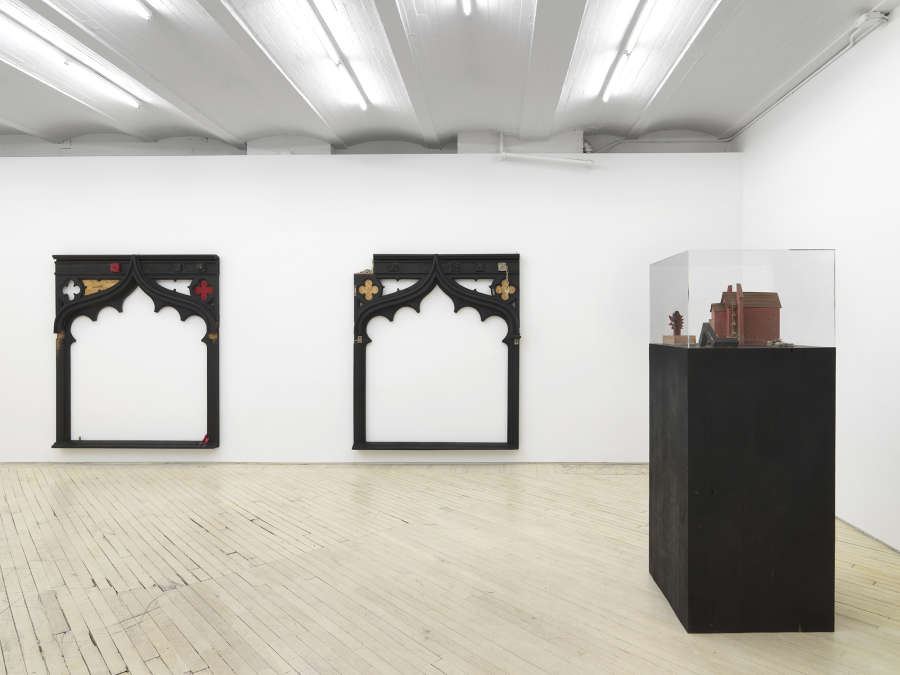 Installation view showing two wooden frame/archway sculptures hanging on the wall, and at right foreground a black plinth with a glass vitrine on top with a model of a red church inside.