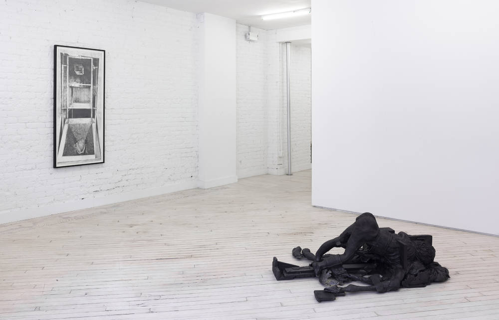 Installation view of the front gallery. At left a black and white graphite drawing framed in black, and at right, in the foreground a black floor sculpture of a figure in a pose with its head touching the ground and rear elevated, combined with what appears to be a decaying horse.