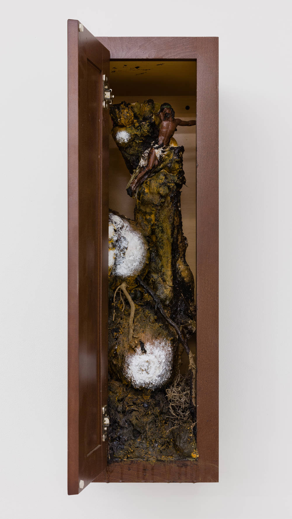 Image of a sculpture by Brandon Ndife. Hanging on a gallery wall is what appears to be a piece of furniture, like a kitchen cabinet, painted brown with dirt, debris and contained growth on the inside of the furniture. What looks to be gourds are "growing" on a central trunk inside of the artwork, painted white like mold. There is a small figurine of a black Jesus in the cabinet.