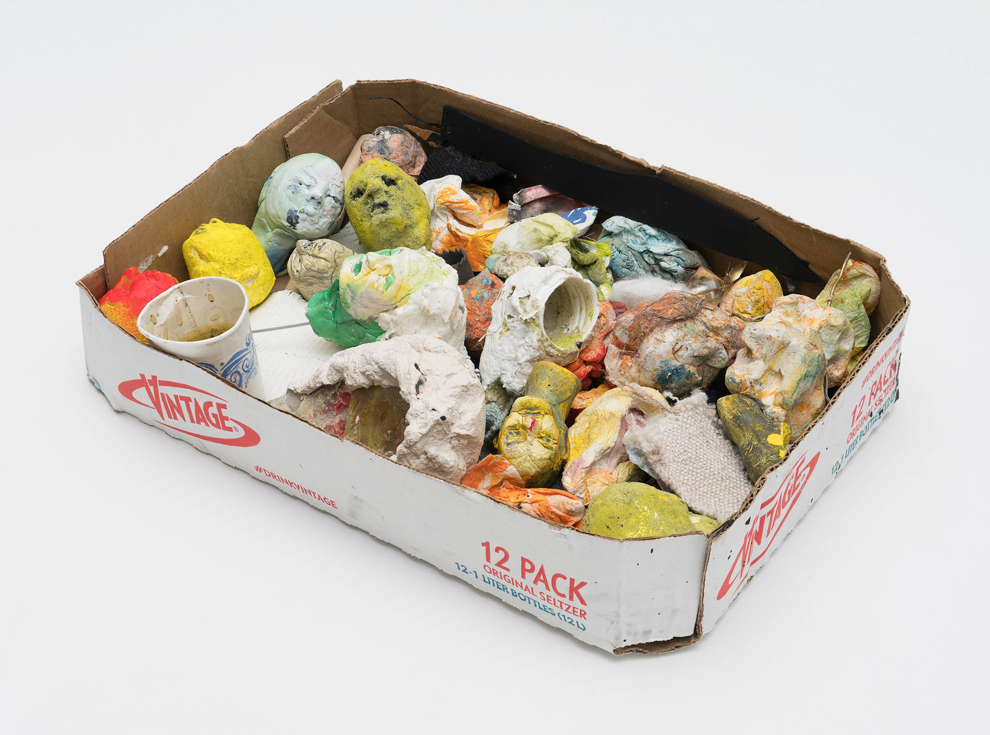 Against a white background, a recycled Vintage seltzer box sits holding various heads from puppets used in the making of films by Tom Thayer.