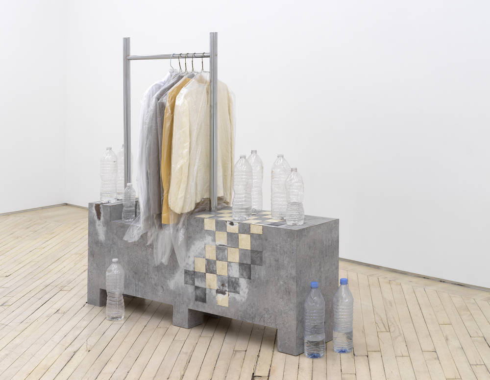 In a gallery space an abstract, architectural sculpture. The object contains a clothing rack housing several pale colored shirts in laundry bags. There is a floor title pattern on the base. Numerous empty water bottles surround the sculpture on the floor and are resting on top of the base.  