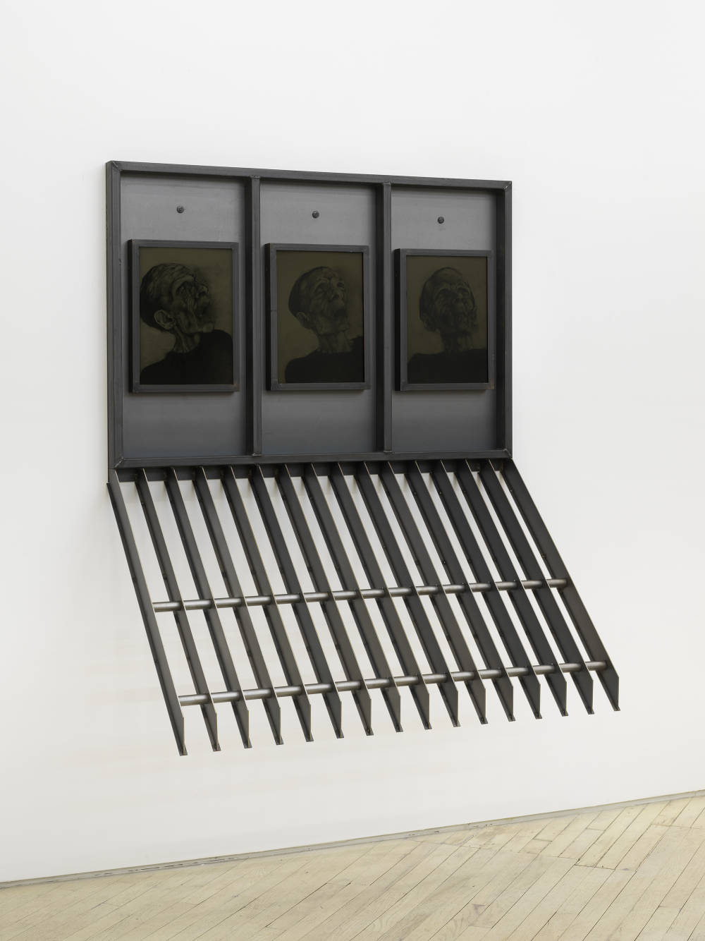 A large steel sculpture hanging from a wall containing three drawings a human head.