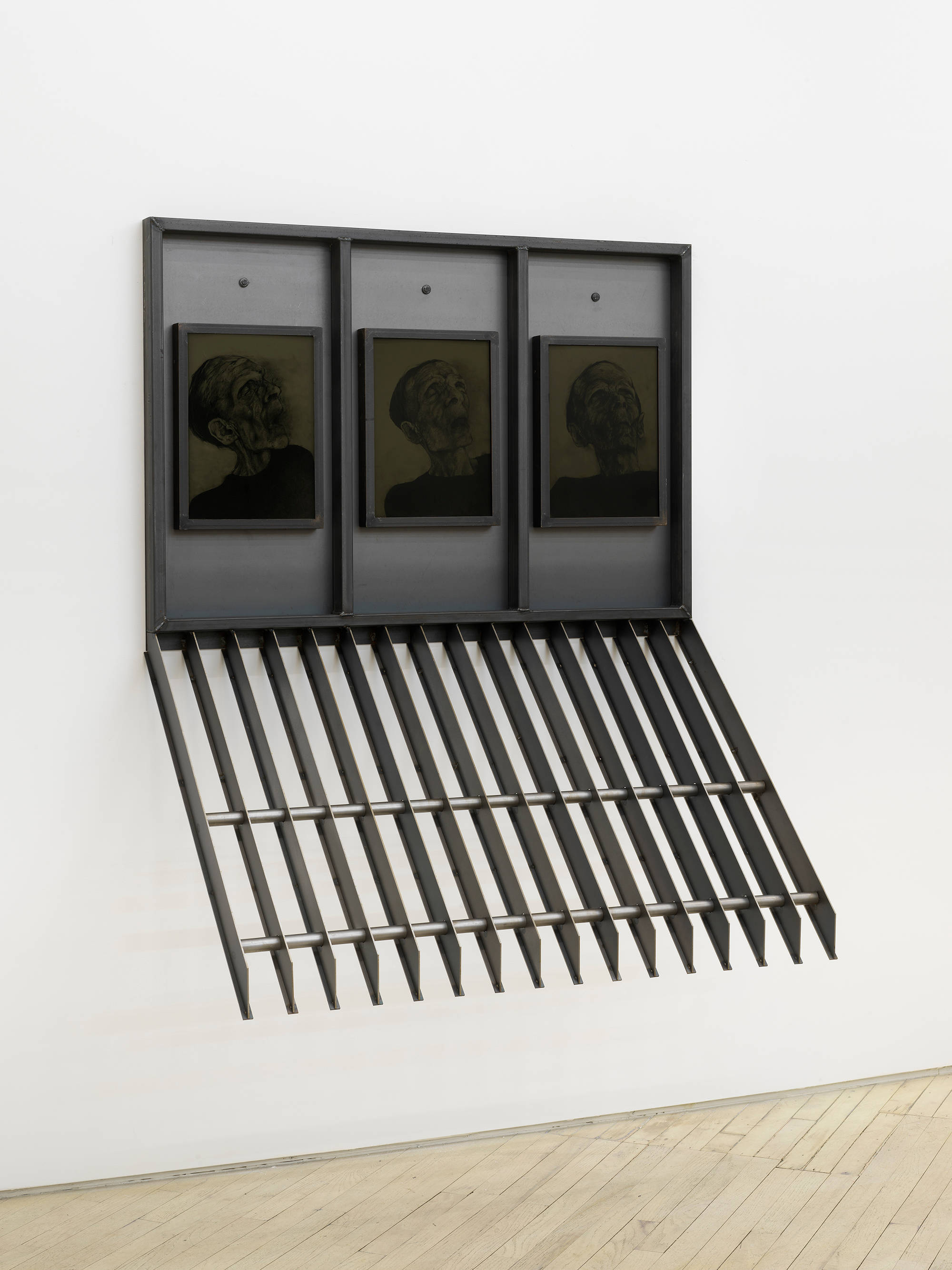 A large steel sculpture hanging from a wall containing three drawings a human head.