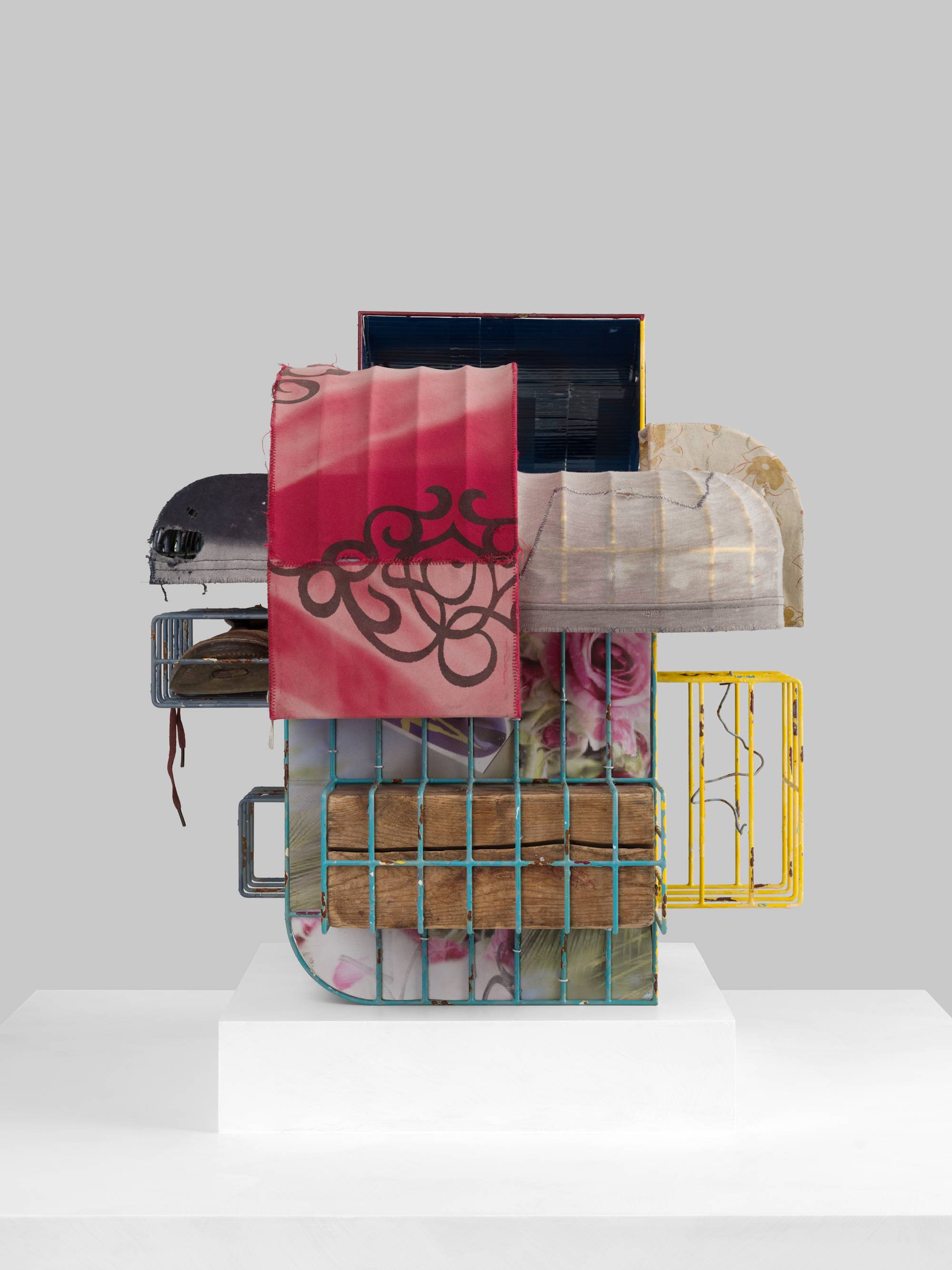 A cage-like colorful steal wire sculpture, wrapped with several tattered fabrics to resemble various awnings, and embellished with an old shoe, a twig, printed coroplast signage, an old blue paint can, a lock, a paper tag, and a wooden block sits on a white pedestal against a white wall.