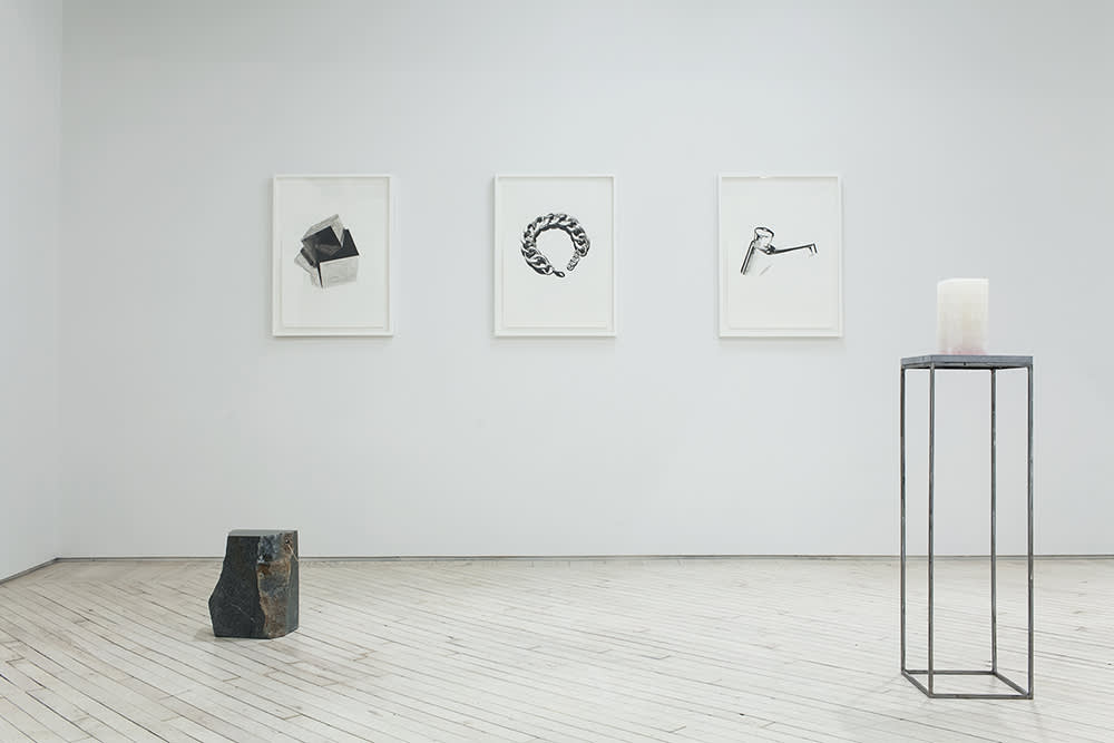 Installation view of 2 sculptures and 3 drawings. At left on the floor is a dark stone with natural edges which has a hole drilled through the top right corner where a large hoop earring pierces the stone, to the right, there is a rectangular plinth made of think steel legs with a blue stone top, on top of it sits a rectangular block of white alabaster, on the bottom is a gradient of pink acrylic paint. The three framed drawings behind on the wall are painted with black ink on white paper and from left depict a piece of the mineral pyrite, a golden bracelet and a typical bathroom faucet.