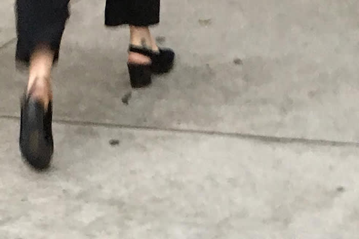 Image of a persons feet and black heeled shoes as they walk away on a side walk.