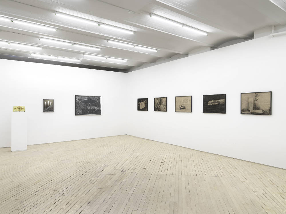 A gallery space with numerous framed objects throughout. On the left wall are two framed black and white photographs or drawings. In the corner is a green vintage radio on top of a pedestal.  On the right wall is a series of pencil drawings behind tinted glass frames.