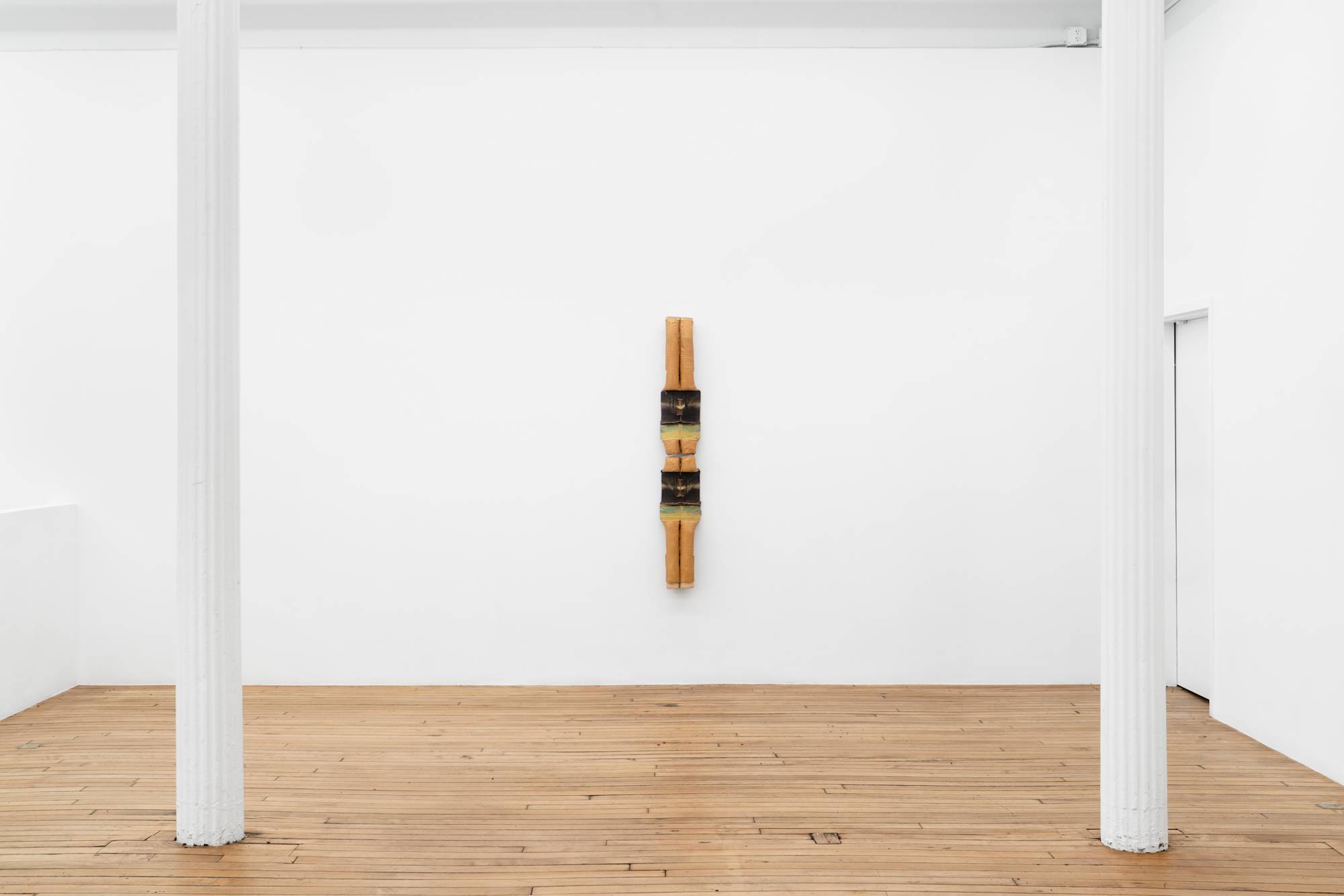 A large, vertically oriented, 2-piece sculpture, featuring cylindrical elements, in hues of mustard, brown, and green, hangs on a white wall, above a wood floor, centered between two white pillars.