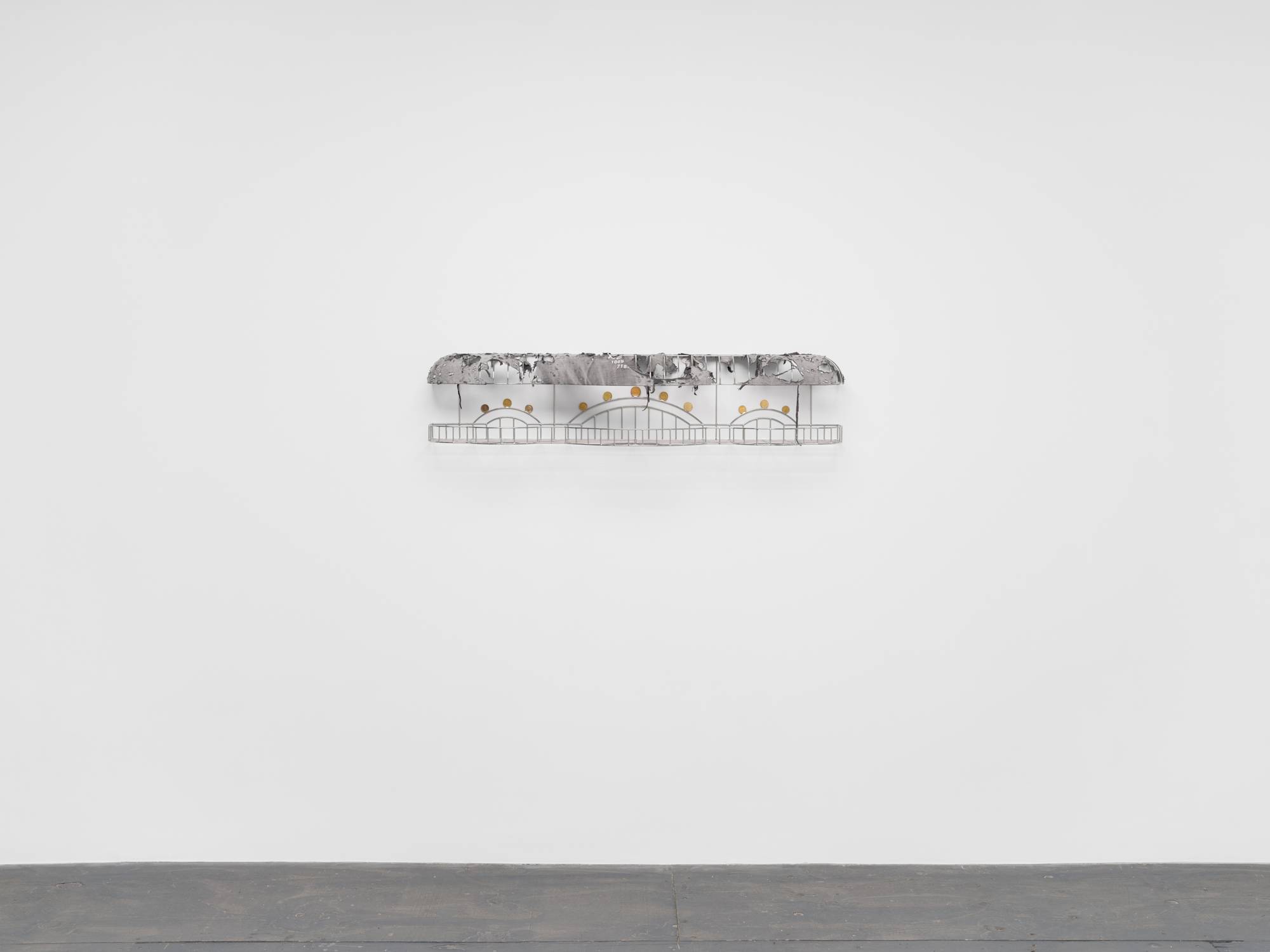A cage-like chrome sculpture, wrapped with a tattered grey t-shirt to resemble an awning, and embellished with soldered on gold-colored coins hangs on a white wall above a grey floor.