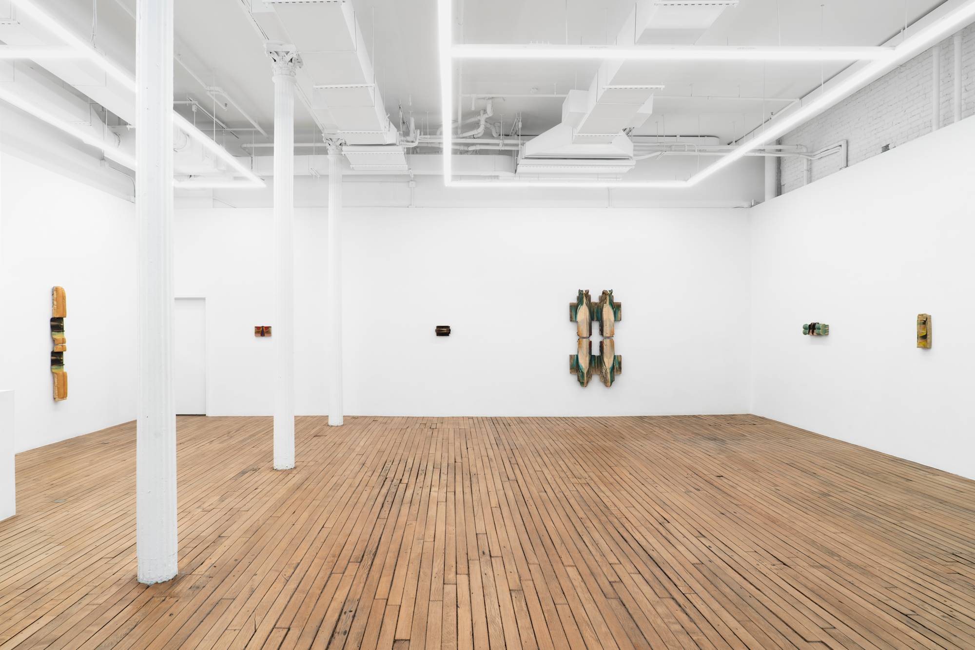 Six wall-mounted clay sculptures of various color and scale hang in a well lit art gallery with white columns and wooden floor.