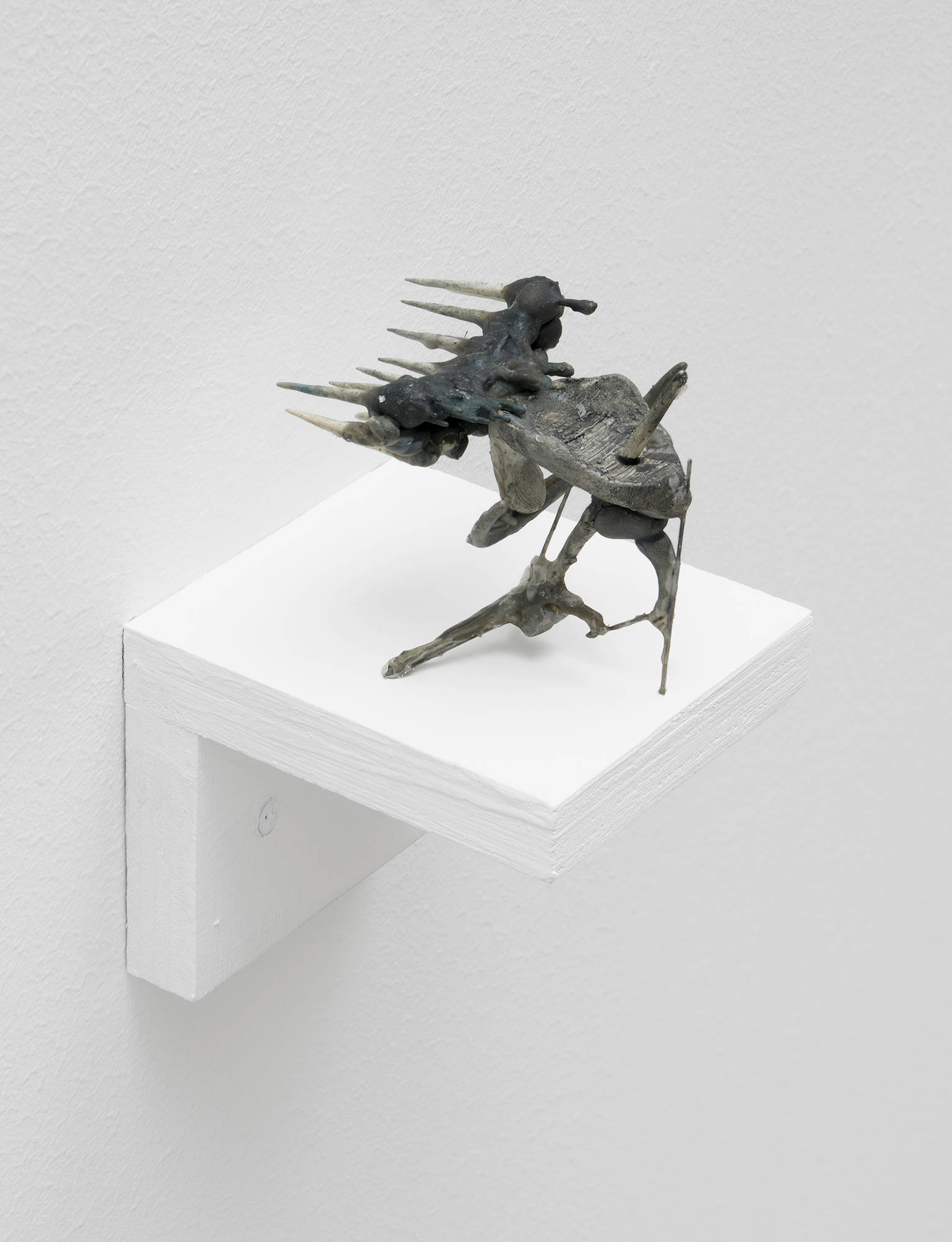 A small, handmade sculpture by Matt Hoyt sits on a white ply wood shelf. It resembles a crude alien space ship sitting on a thin stand, depicted in dark shades of green and grey.