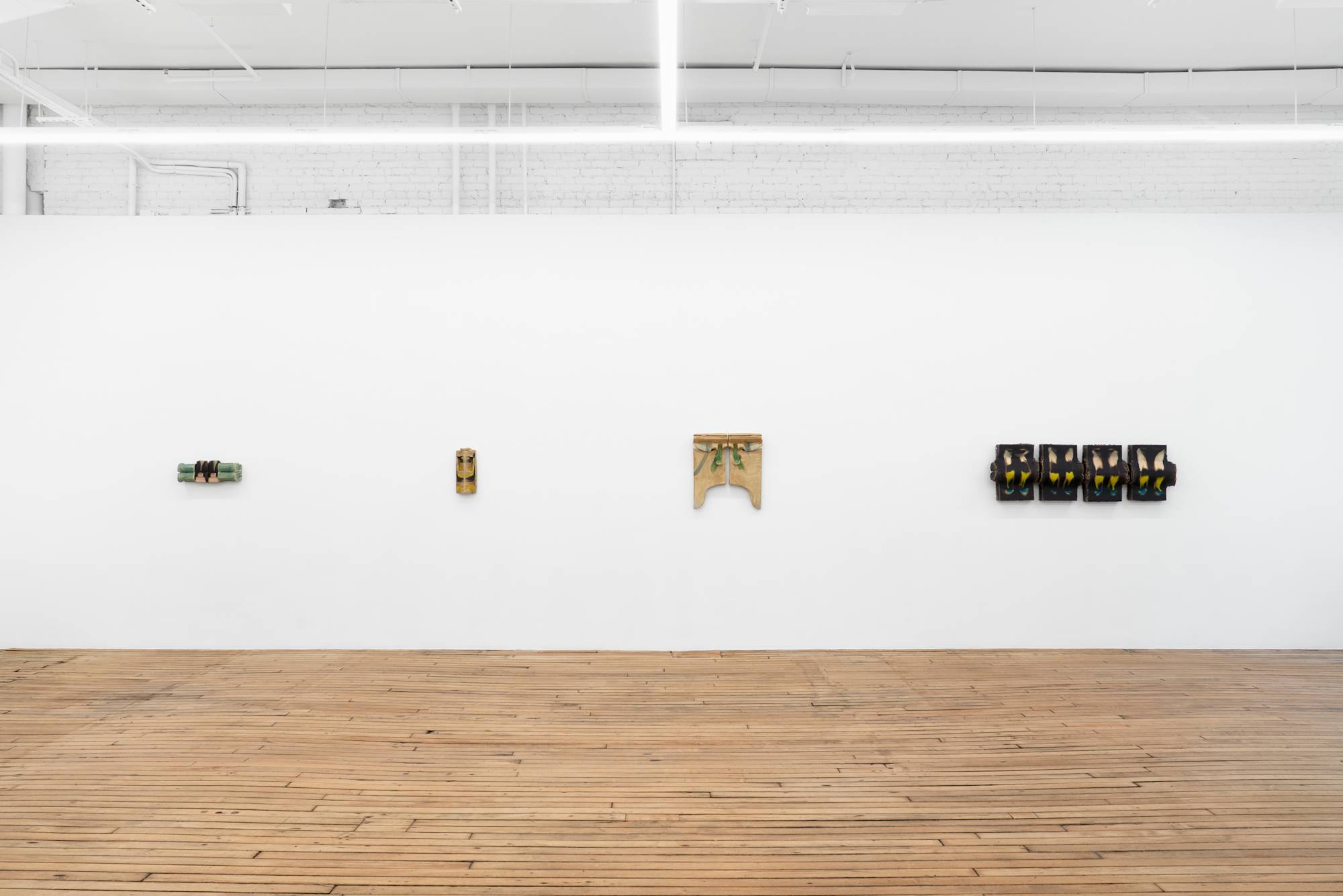 Four wall-mounted ceramic sculptures of various color and scale hang in a line on a white wall in an art gallery with a wooden floor.