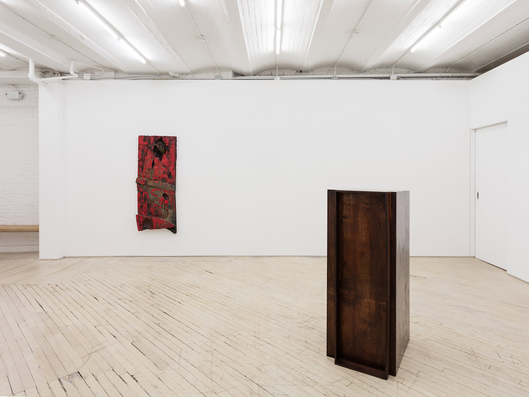 Installation view of an exhibition by Brandon Ndife. In a gallery there is sculpture sitting on the floor appearing like pieces of furniture or cabinetry, stained brown. Hanging on the wall is another sculpture that appears to look like a dirty red door.