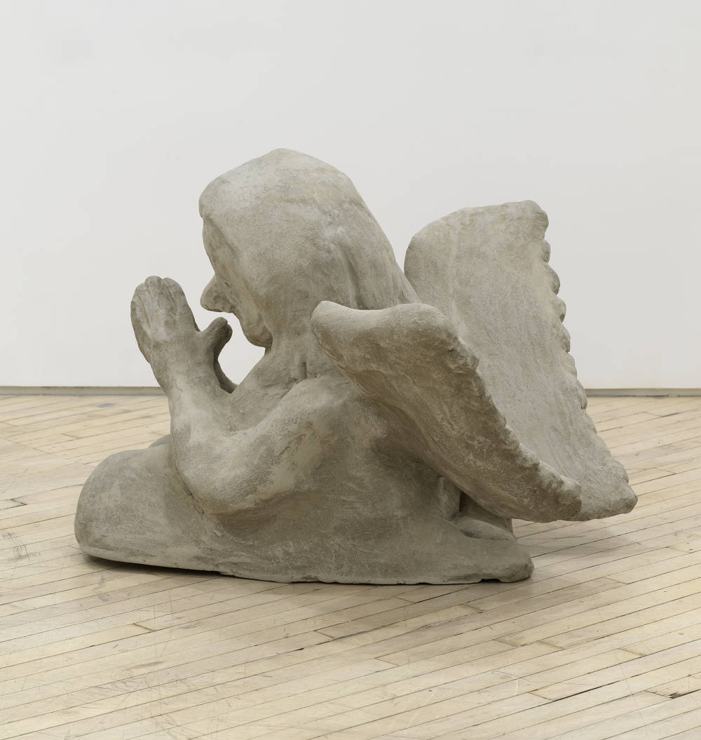 Image of an artwork by Libby Rothfeld of a cartoonishly rendered angel seated on the floor with wings and hands in a prayer like pose. The angel is sculpted from concrete.