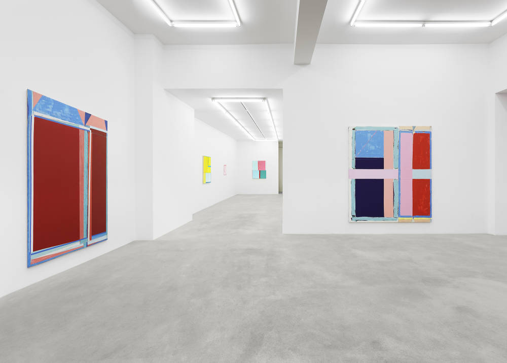 In a gallery space, two large abstract paintings hung on separate walls. The paintings are formally dominated by large rectangular blocks of color. The most striking colors in both paintings are a range of reds, pinks, and blues. The paint is very thick and the canvas is rough. In an additional room in the background are three smaller paintings that are yellow, pink, and green.