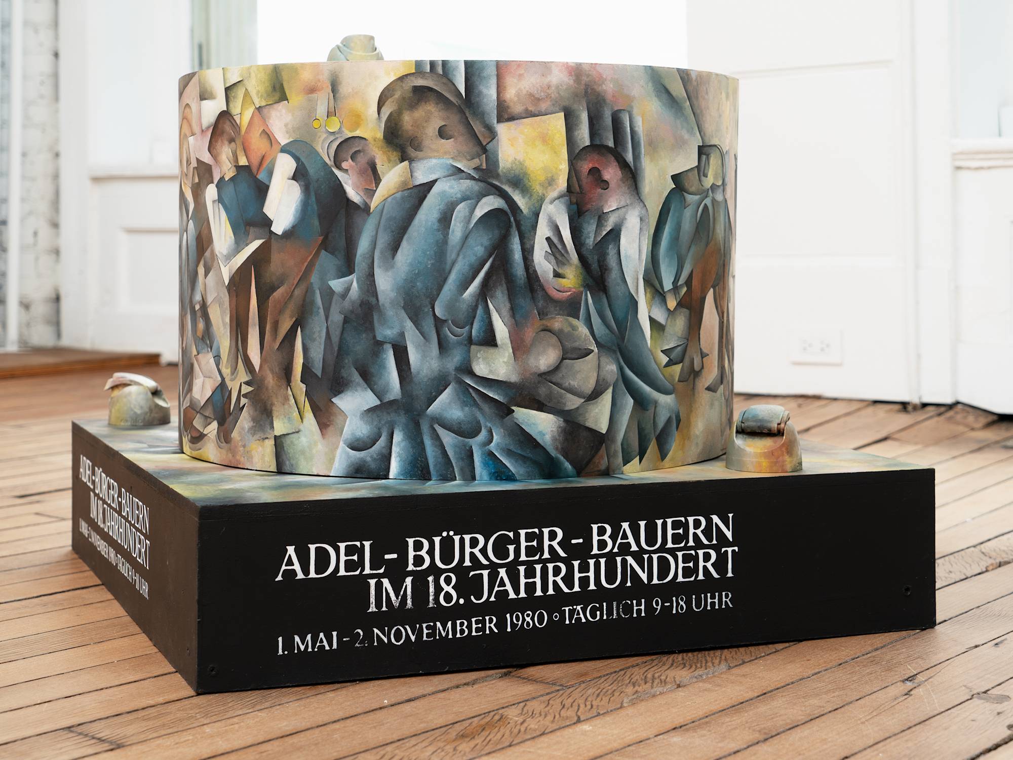 A freestanding sculpture with a round center sits in a gallery space in front of a window. The sculpture is painted in a Cubist style.