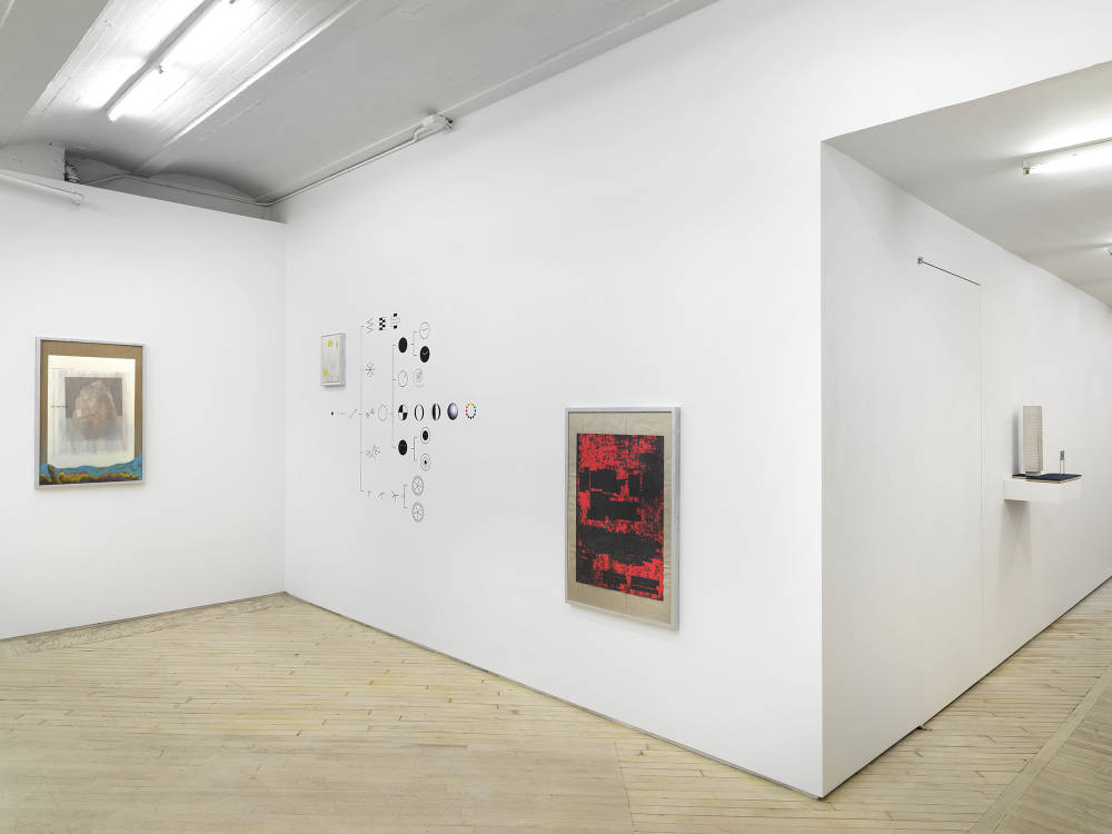 In the corner of a gallery space, a group of three abstract images are installed in frames at varying heights. In the center of the wall is a vinyl installation. 