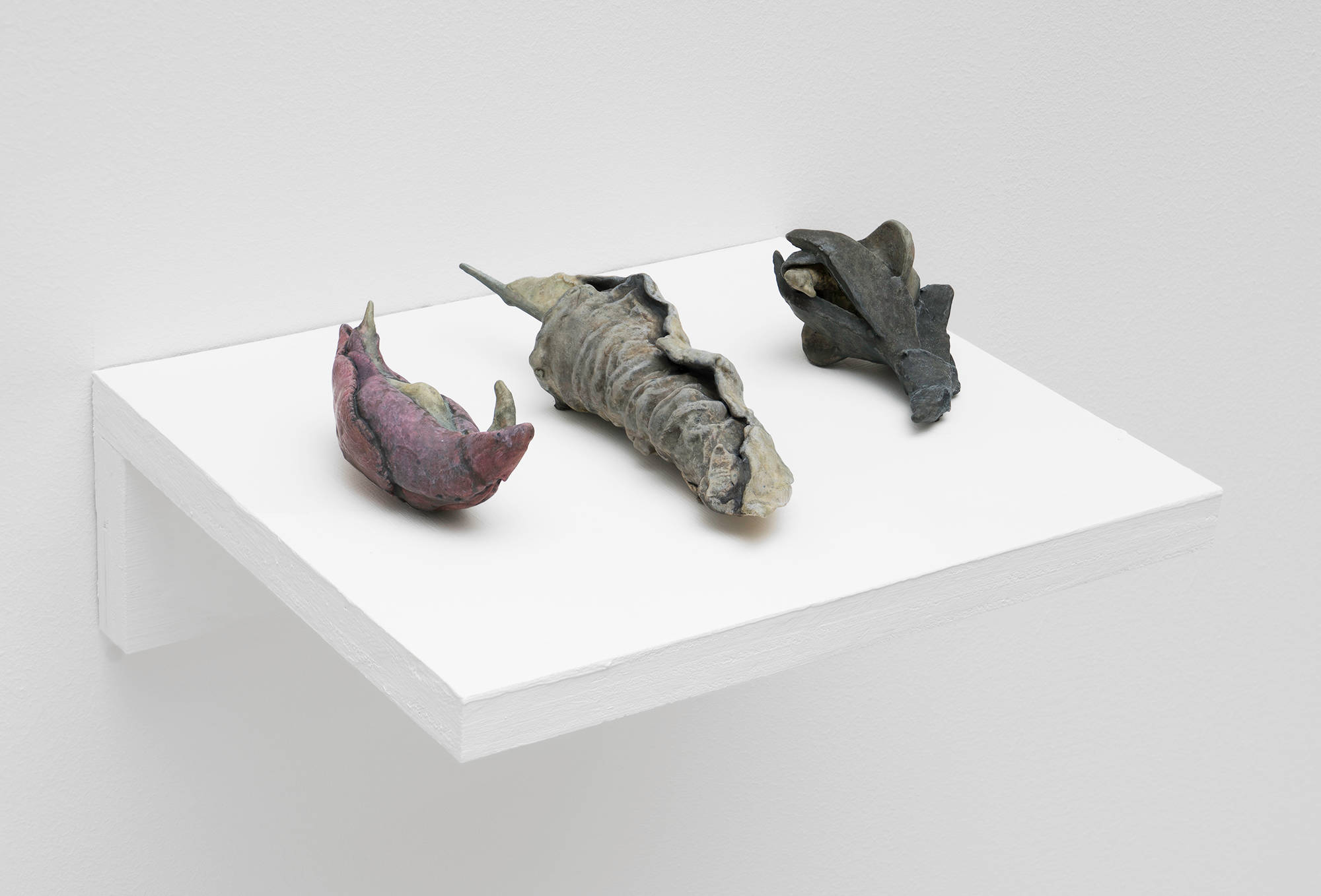 A trio of small, yonic sculptures by Matt Hoyt sit on a white plywood shelf. They are grey, black, and pink in color.