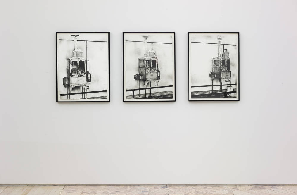 Installation view of 3 framed graphite drawings on a wall. Each one shows a rendering of a rectangular contraption with small doors with windows and legs that keep it off the ground and a pipe attached to the ceiling. 