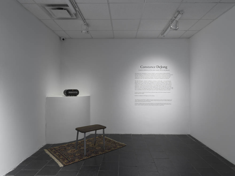 In the corner of a gallery space a vintage radio is placed on a shelf with a small bench resting on top of a carpet. To the right is a wall text. 