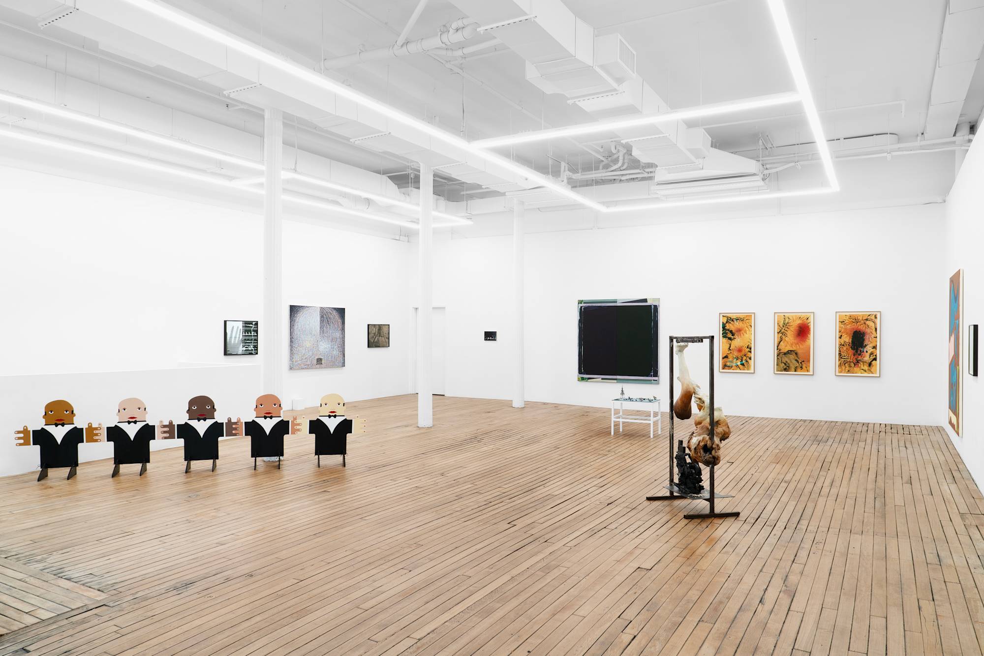 A mix of paintings, drawings, and sculptures fill the walls and floor of a brightly lit gallery with white walls wood floors.