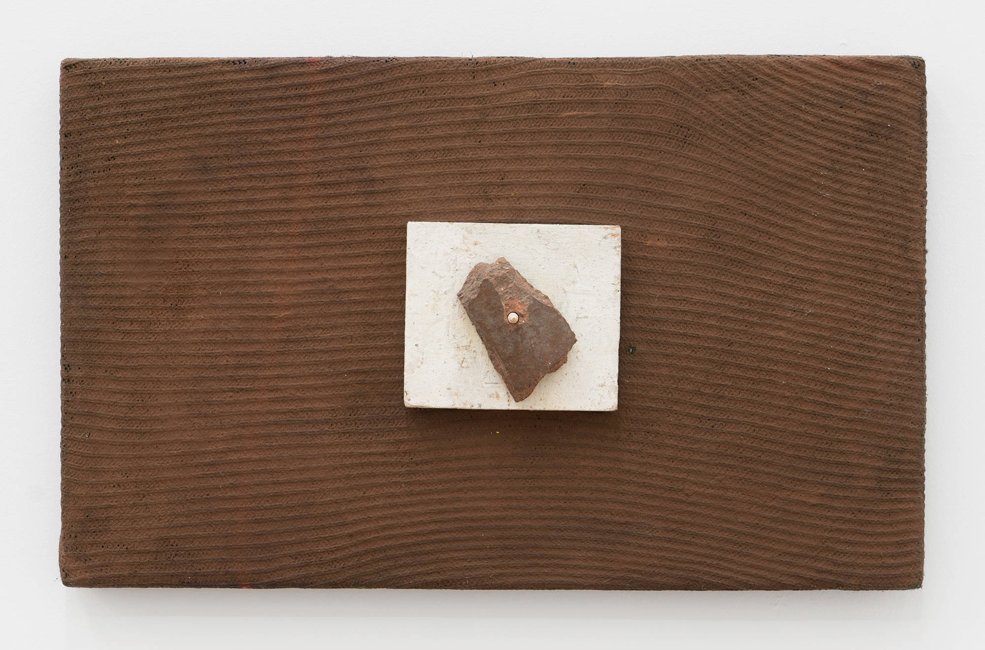 A brown painting on textured fabric. A white square of wood with a spike is secured to the surface of the painting. A brown rock sits impaled on the spike.