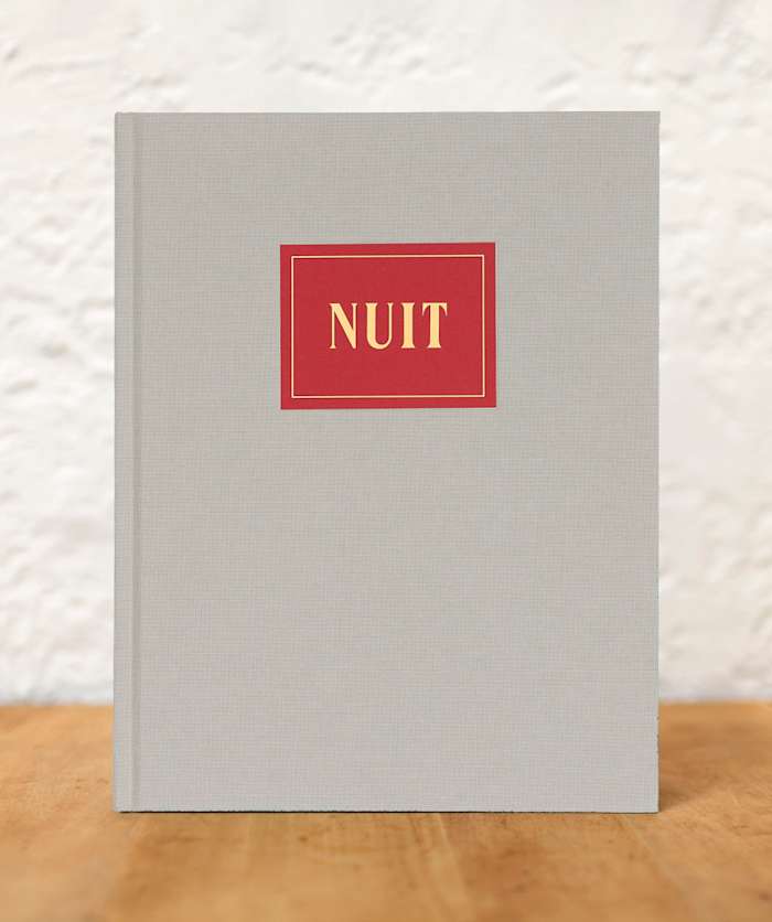A book wrapped in grey cloth with the title "NUIT", spelled in capital letters and gold font, set on a dark red card with thin gold border. The book is standing on a blurry wood table against a white wall.